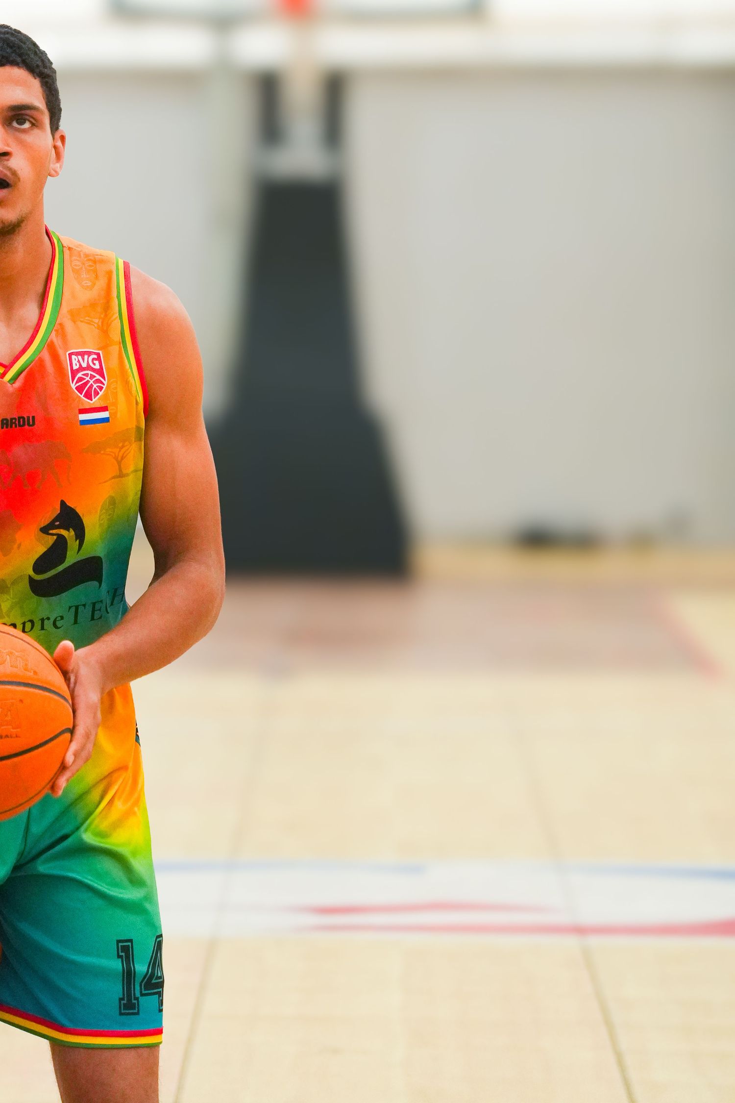 Young male basketball player in colorful rainbow jersey number 14 holding a basketball on an indoor court.