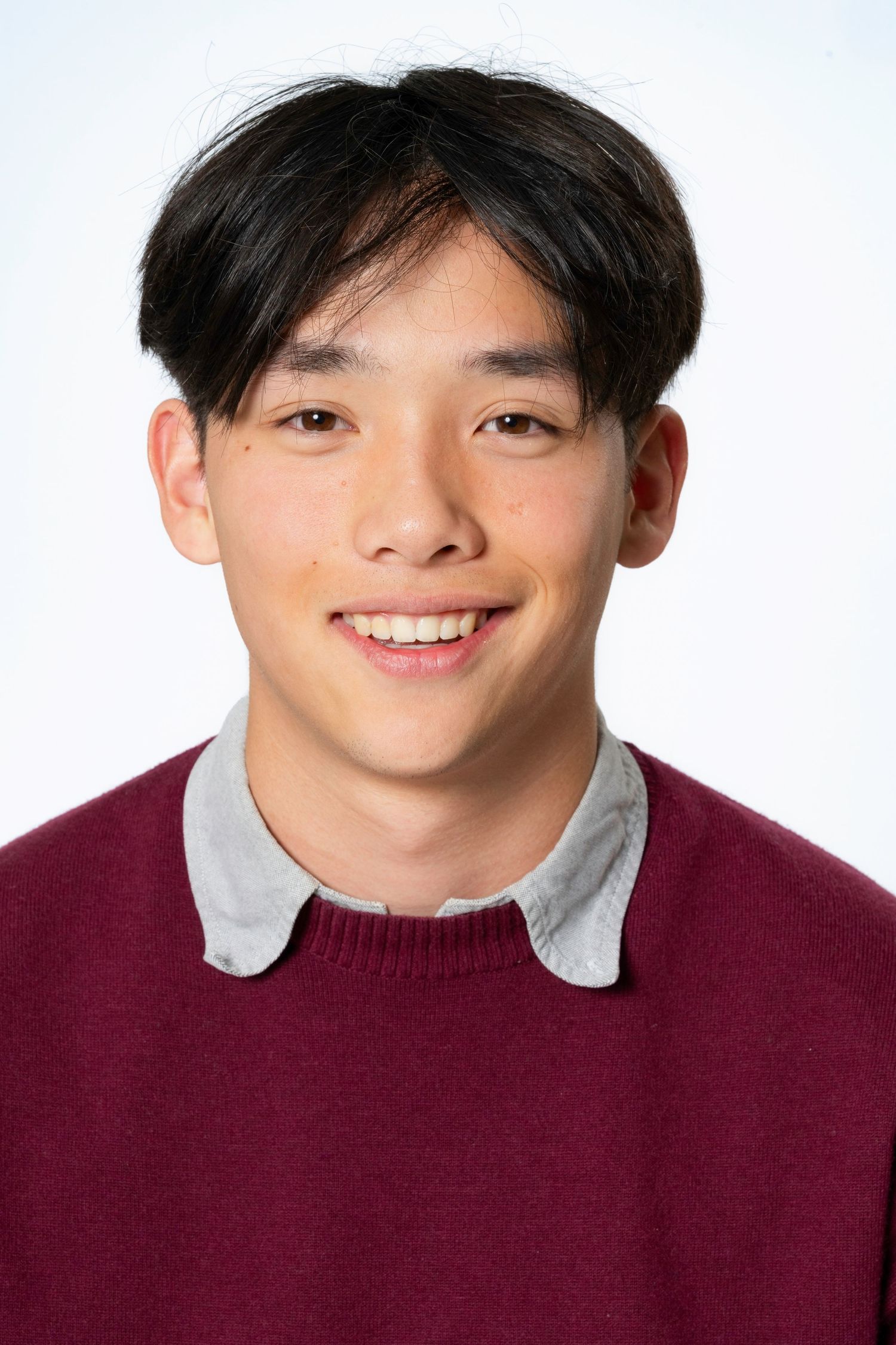 Smiling young man with short dark hair wearing a maroon sweater over a gray collared shirt against a white background.