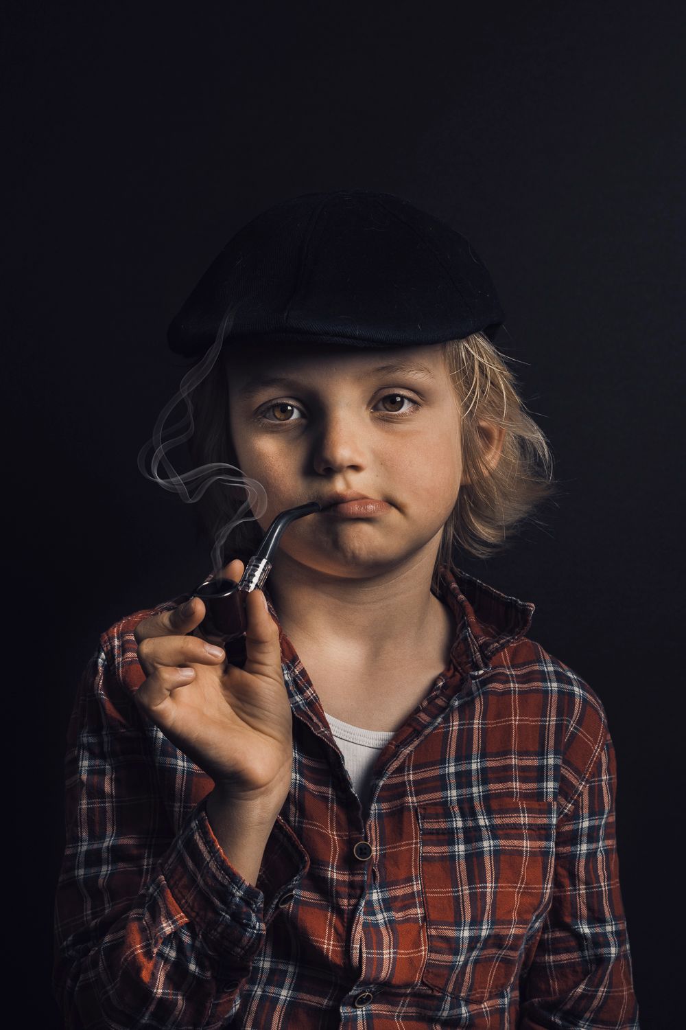 Vintage-style portrait of a child wearing a cap and plaid shirt while holding a pipe.