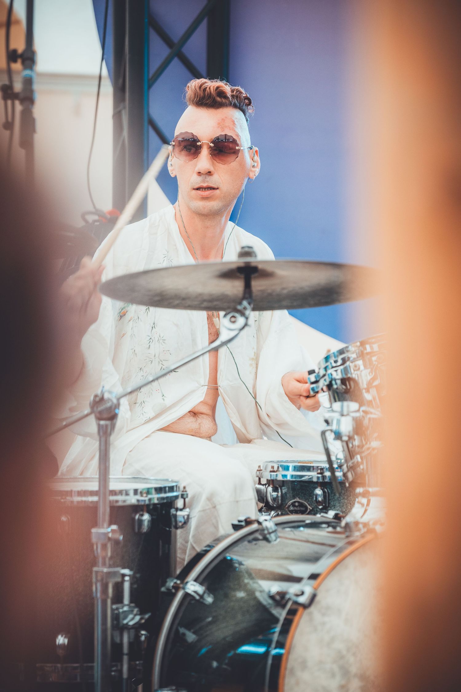 A drummer wearing sunglasses and white clothing performs on a drum kit during a live performance.