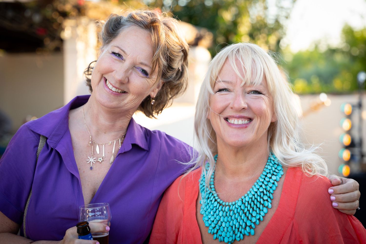 Two women wearing purple and coral colored tops smile warmly while enjoying drinks outdoors on a sunny day.