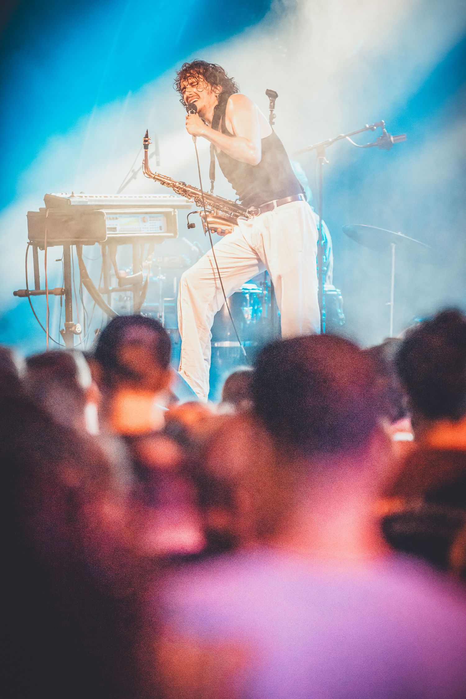 A performer on stage holding a microphone stands energetically in white pants as blue lights shine and crowd watches from below.