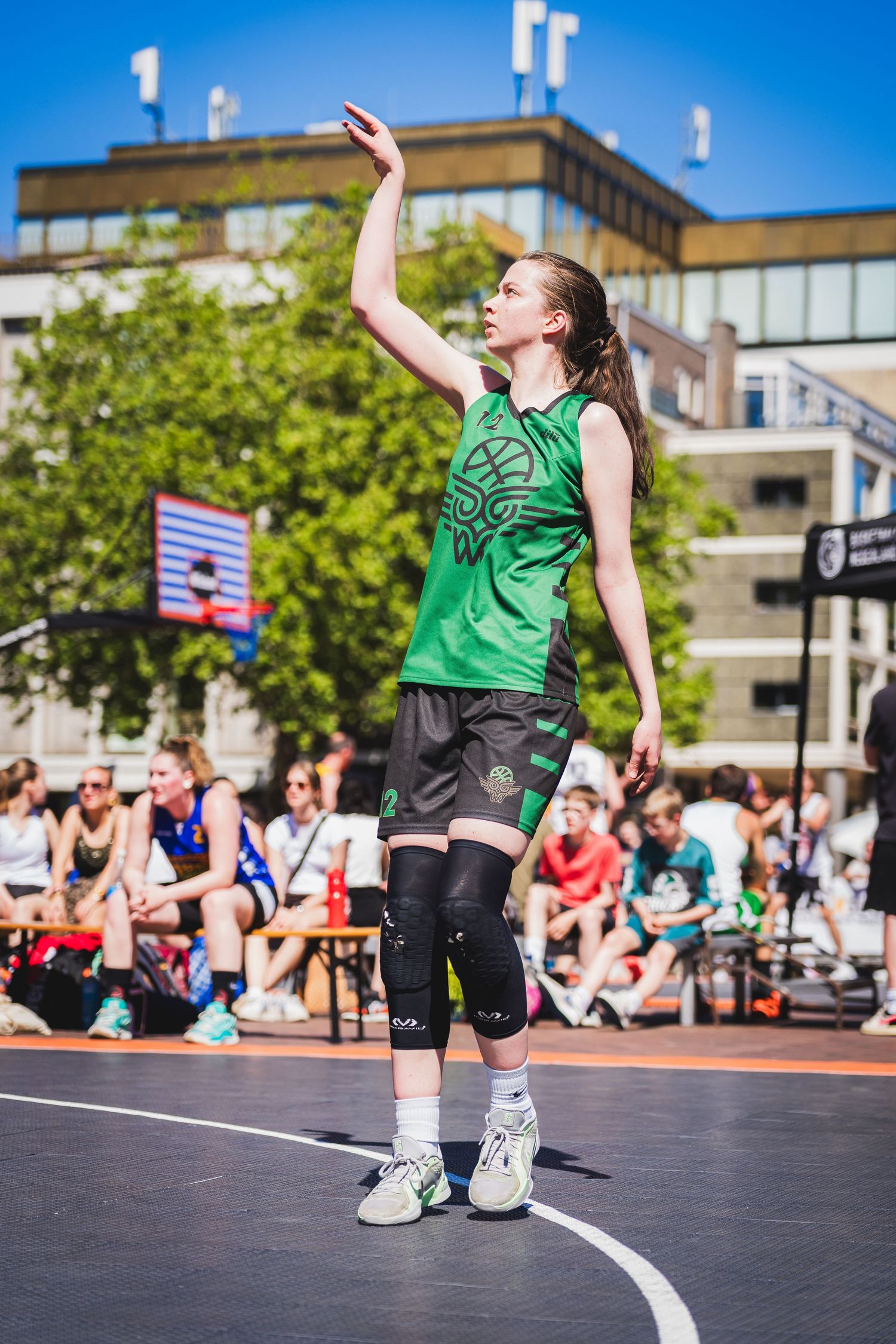 A young female basketball player in a green jersey follows through on a shot during an outdoor 3x3 street game.