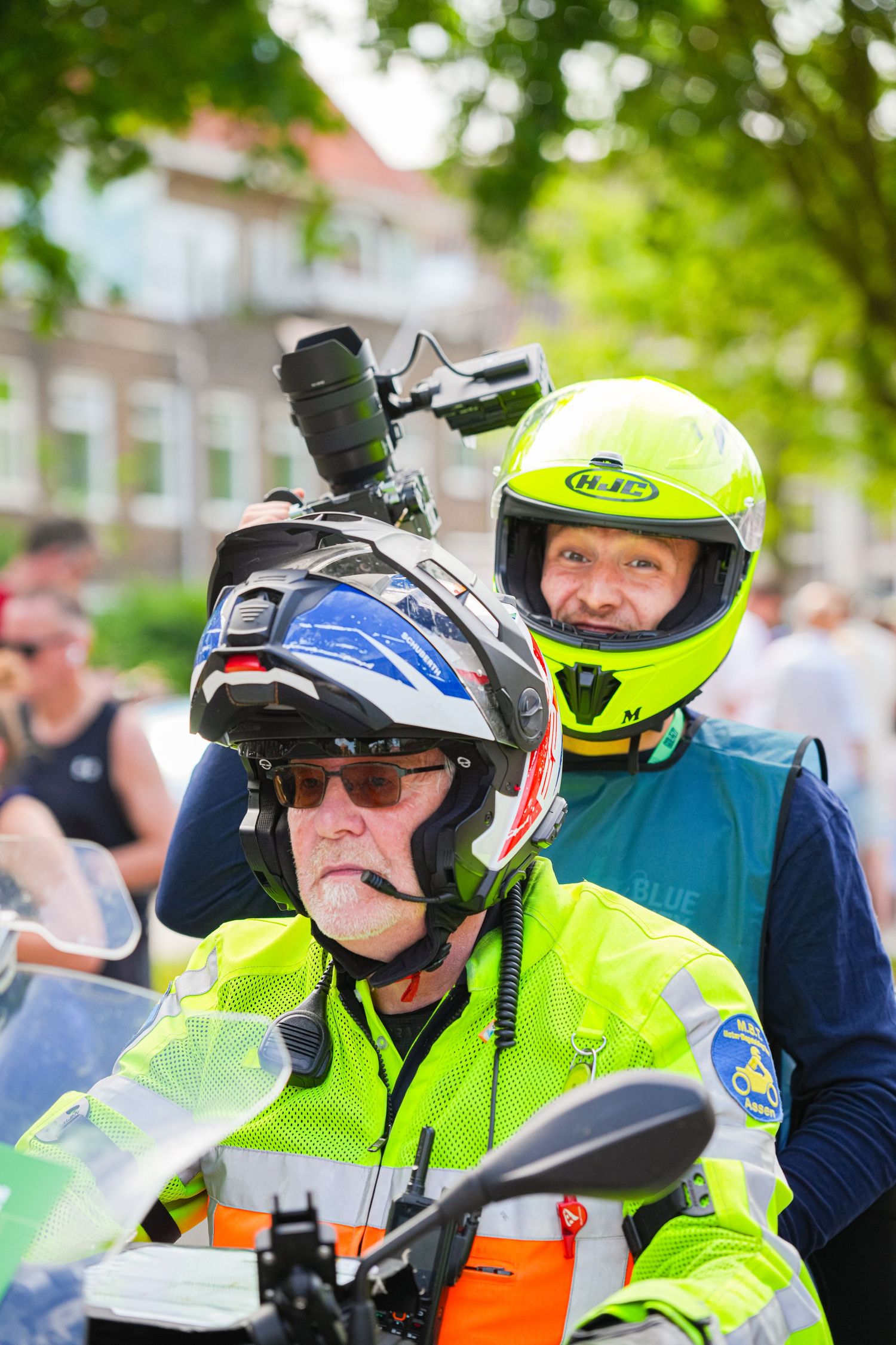 Two motorcycle riders wearing bright yellow helmets and safety vests, with a camera mounted on helmet, at outdoor event.