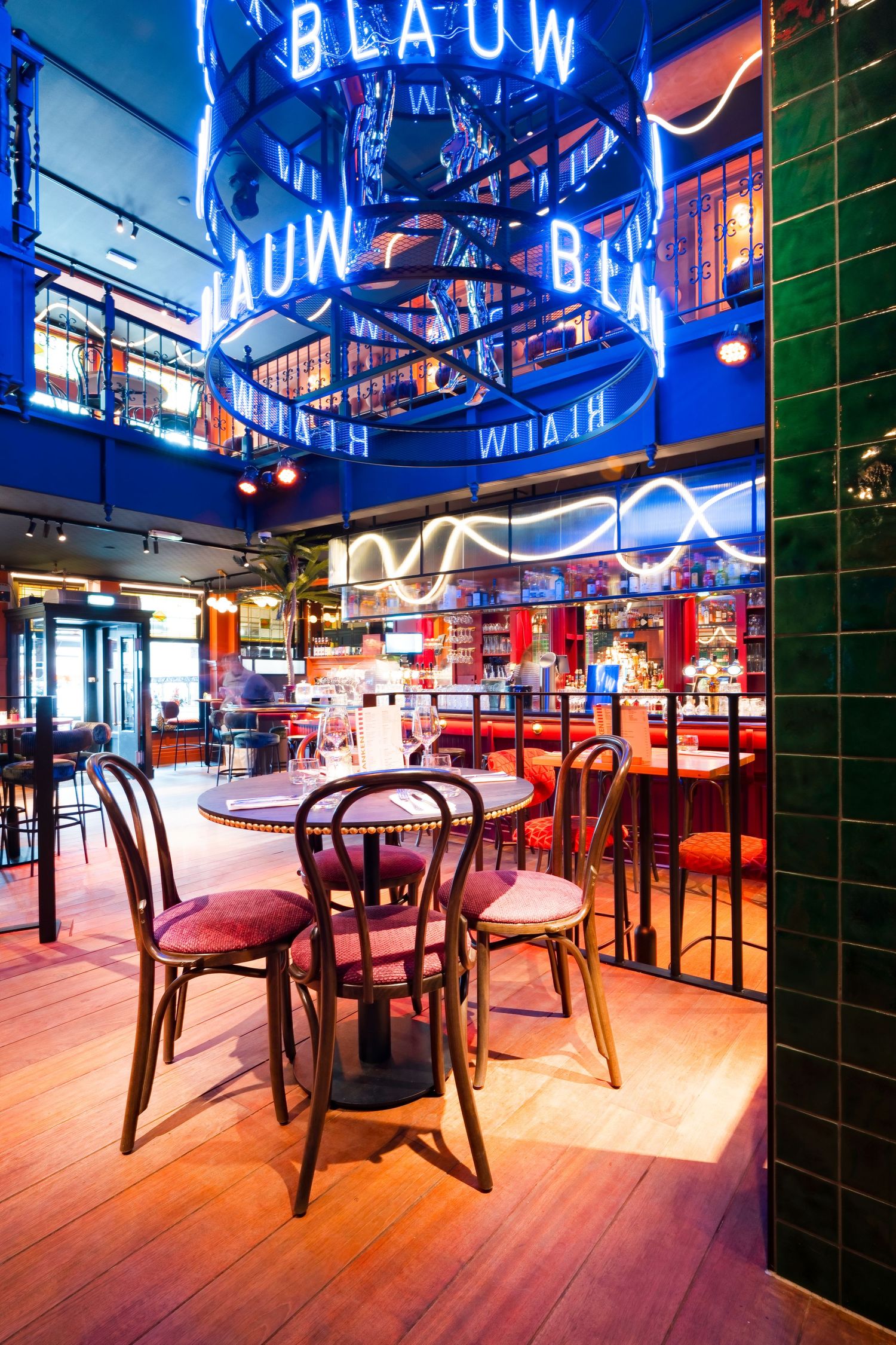 Vibrant restaurant interior with neon BLAUW signs, wooden bistro chairs, round tables, and green tiled walls under colorful lighting.