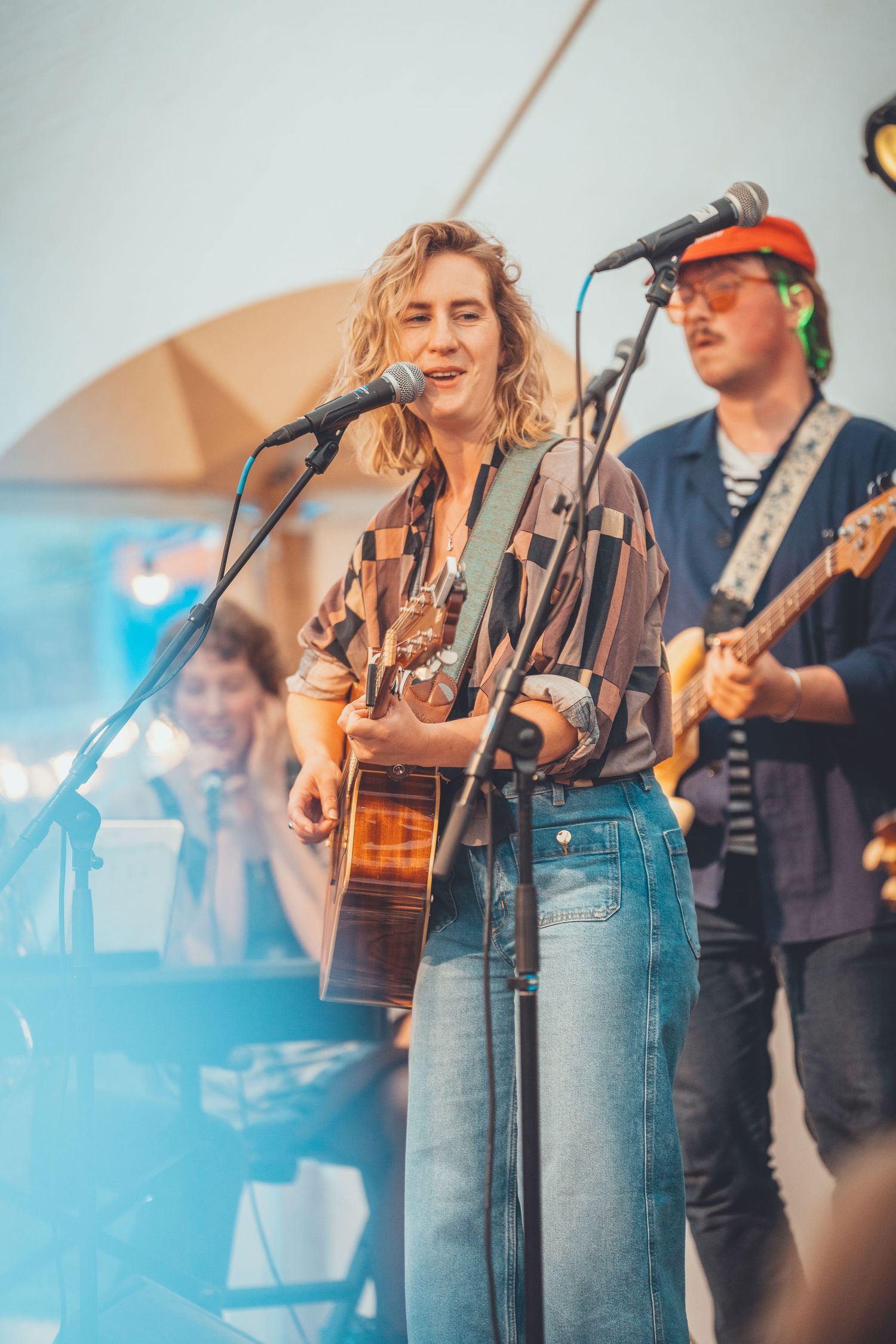 Musicians perform on stage with acoustic guitars and microphones at an outdoor concert venue in blue lighting atmospheres.