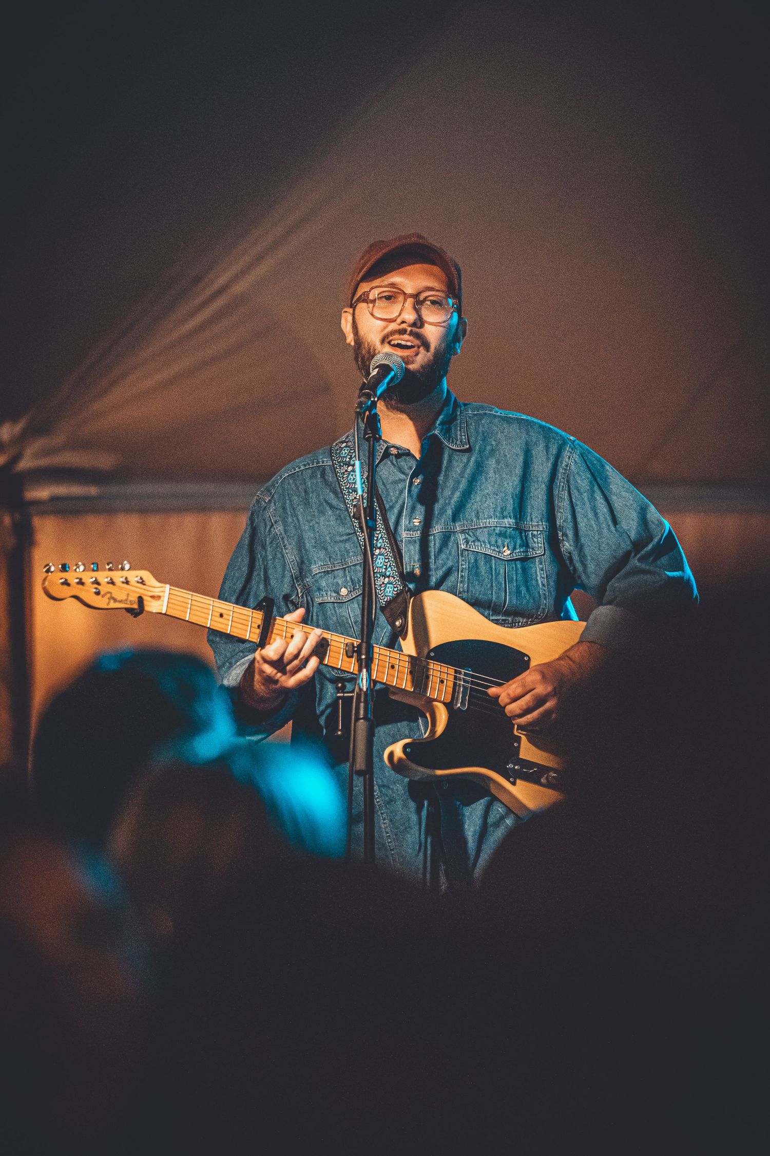 A musician in denim jacket performs on stage with a guitar under warm lighting at a concert venue.