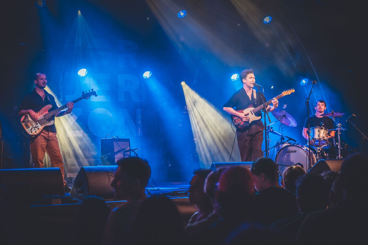 A band performs on stage with dramatic blue lighting and fog effects while playing guitars and drums during a live concert.