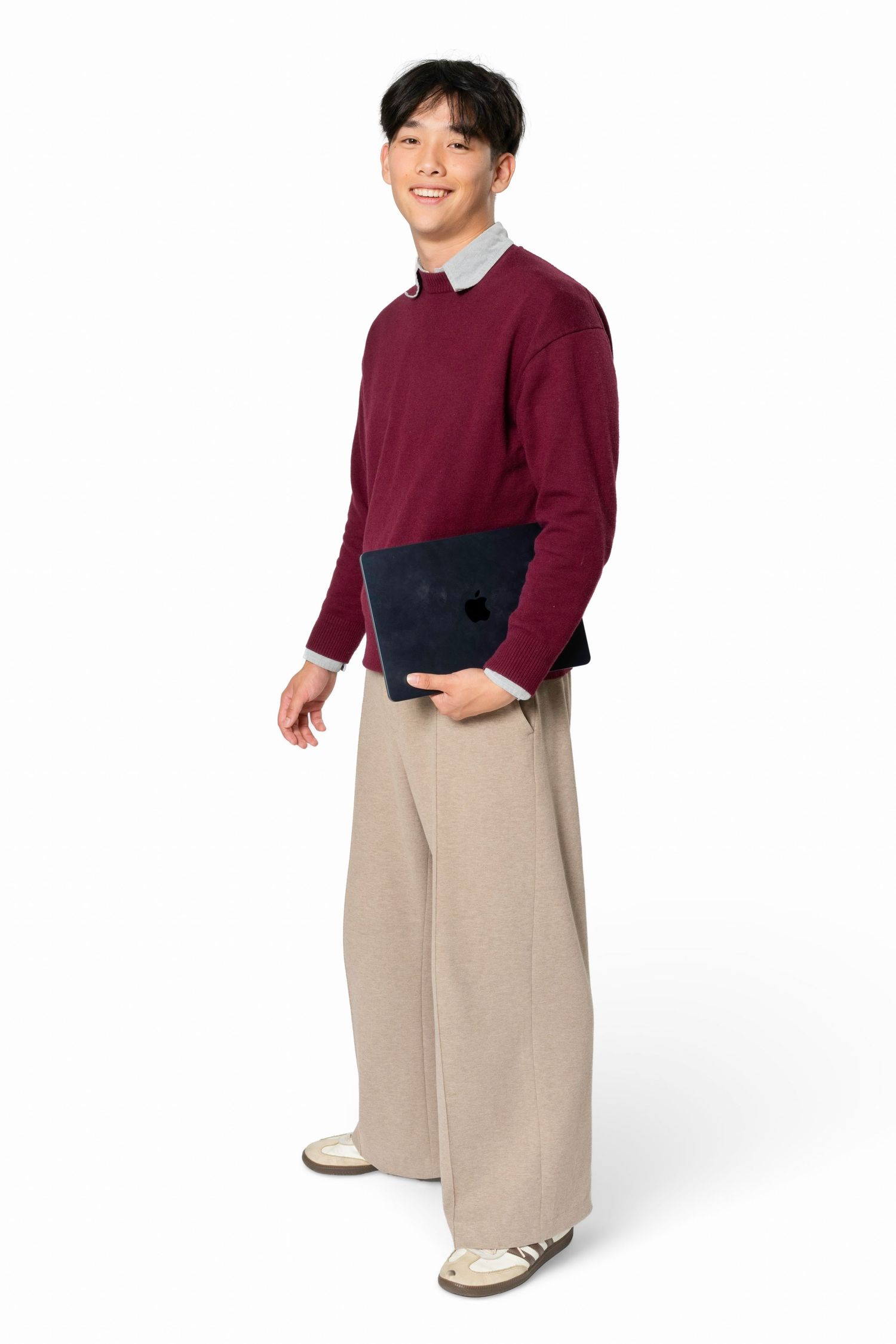 Smiling young man in a burgundy sweater over a gray collared shirt and wide beige trousers holds a dark laptop against a white background.