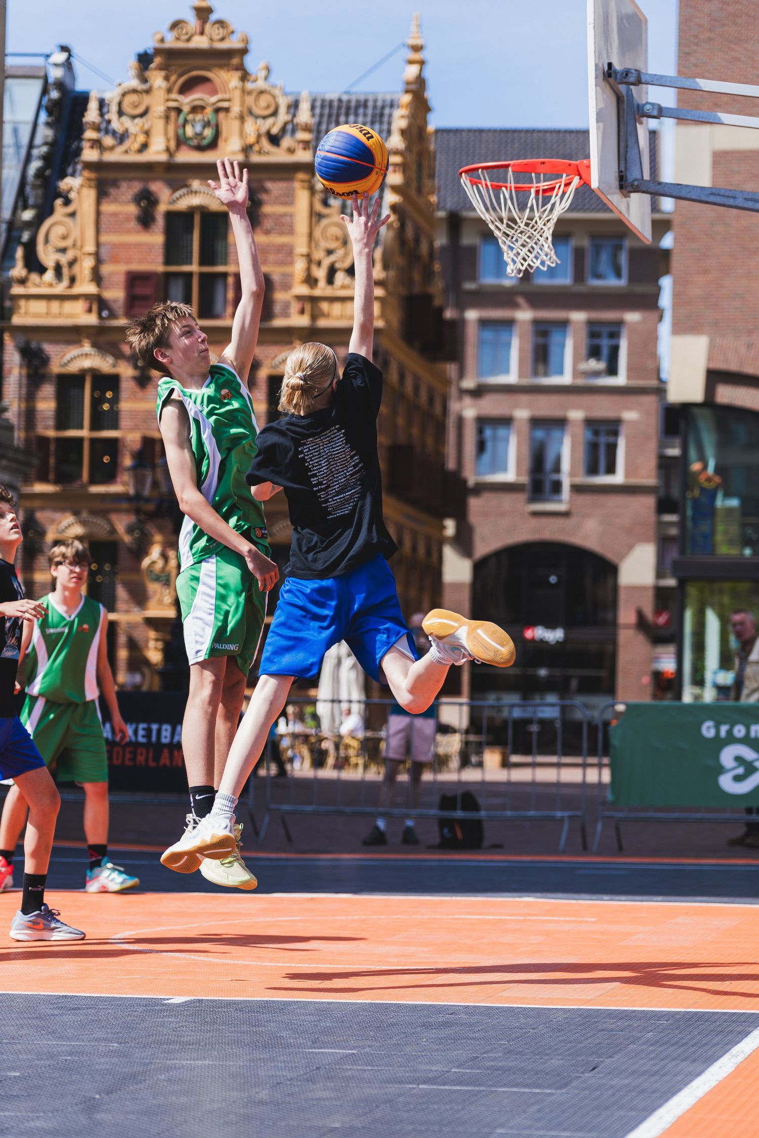 Young players compete in an outdoor 3x3 basketball tournament on an orange court with historic European buildings in background.