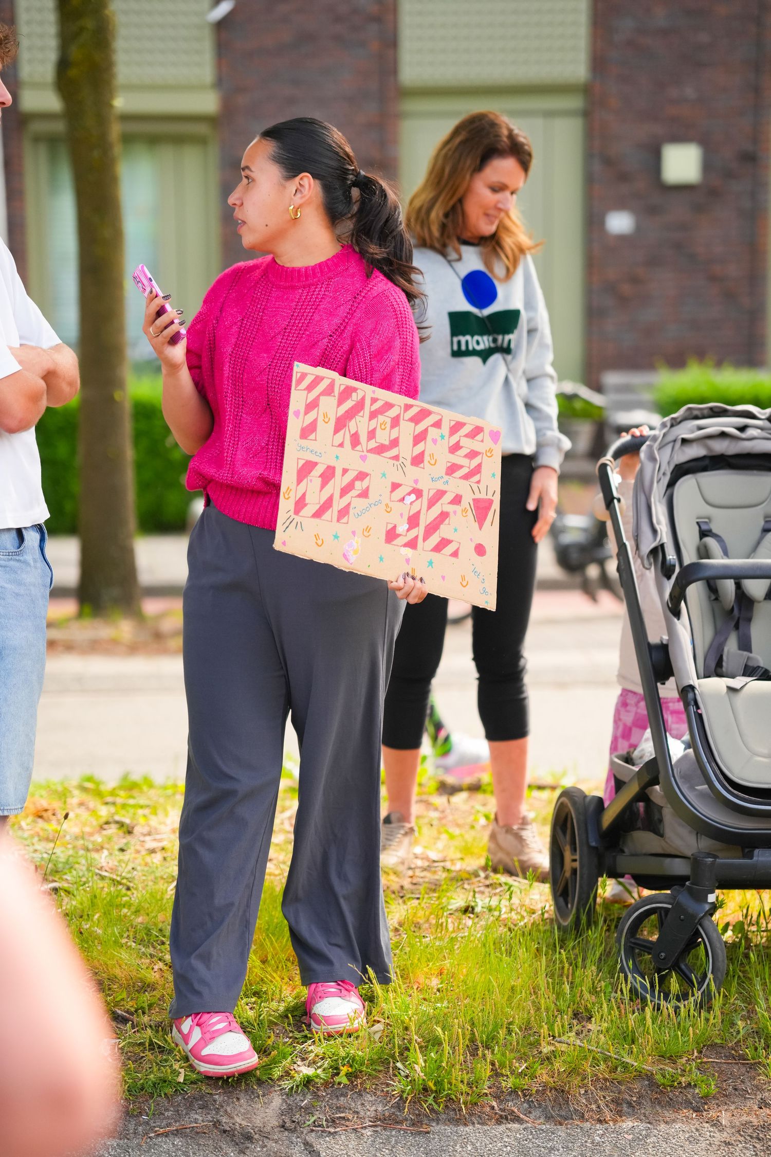 Woman in pink sweater holds protest sign while checking phone at outdoor rally with stroller and crowd nearby.
