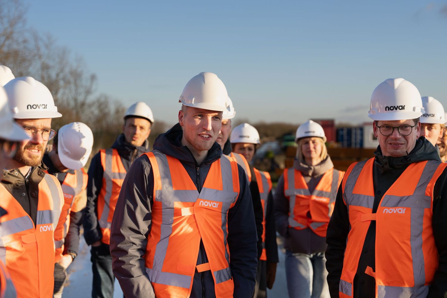 Group of people wearing orange safety vests and white hard hats at a construction site on a sunny day.