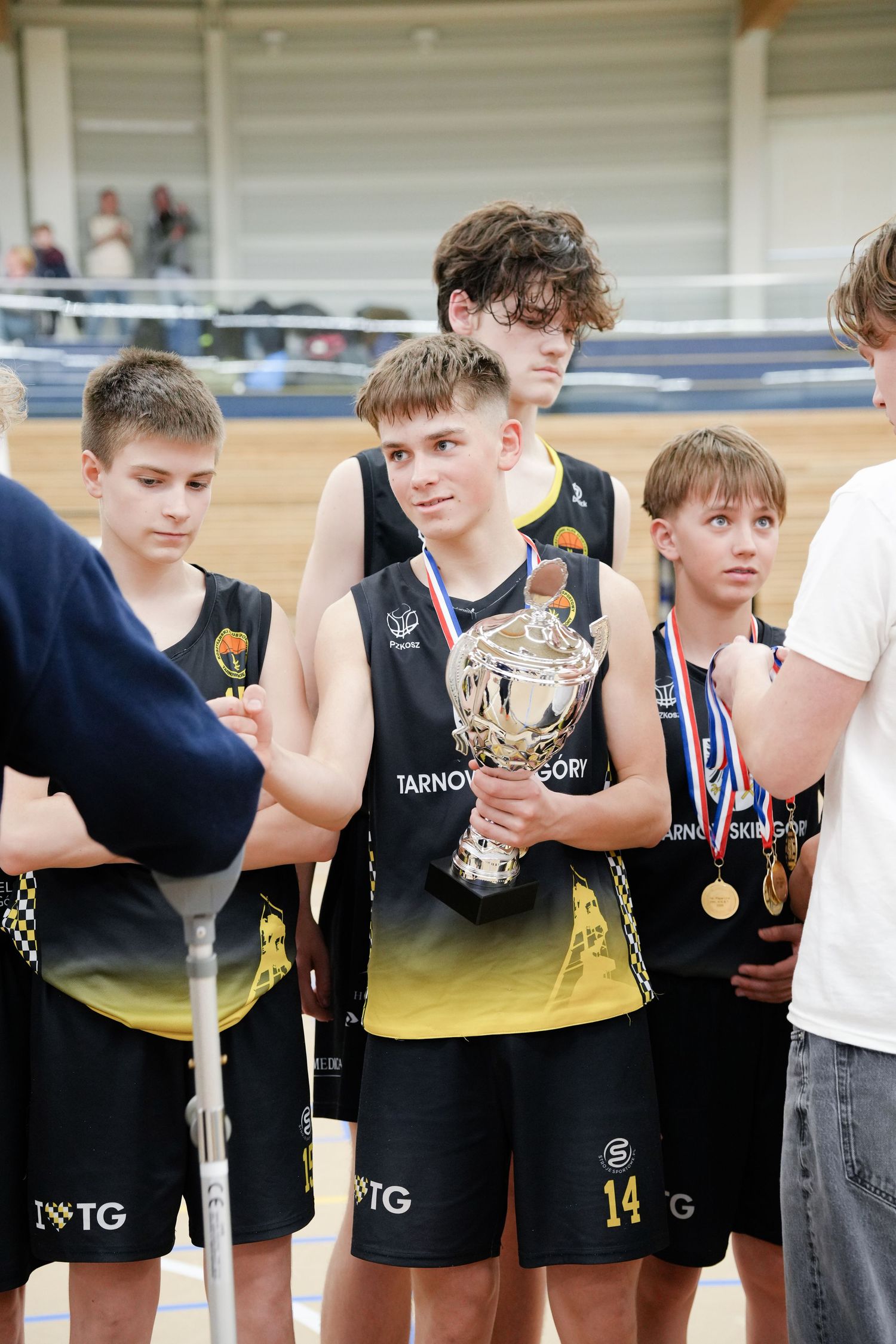 Young basketball players in black and gold uniforms holding a trophy and medals at an indoor sports award ceremony.