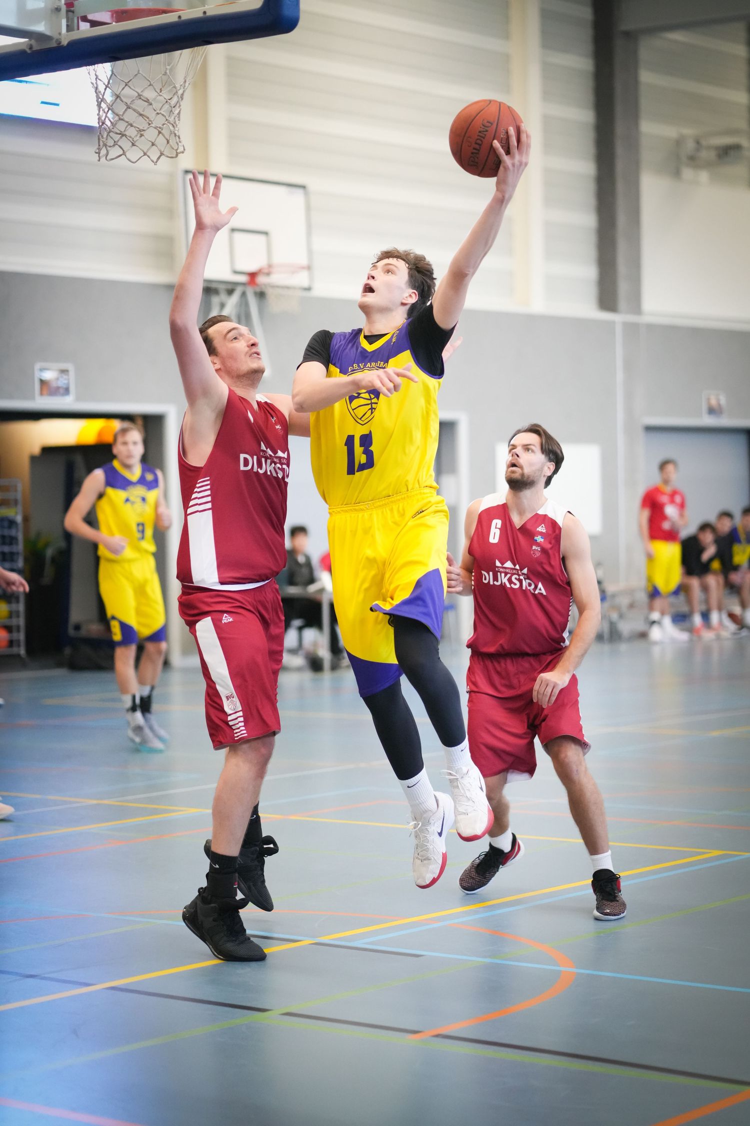Basketball player in yellow jersey drives to the basket against defenders in red jerseys during indoor game.