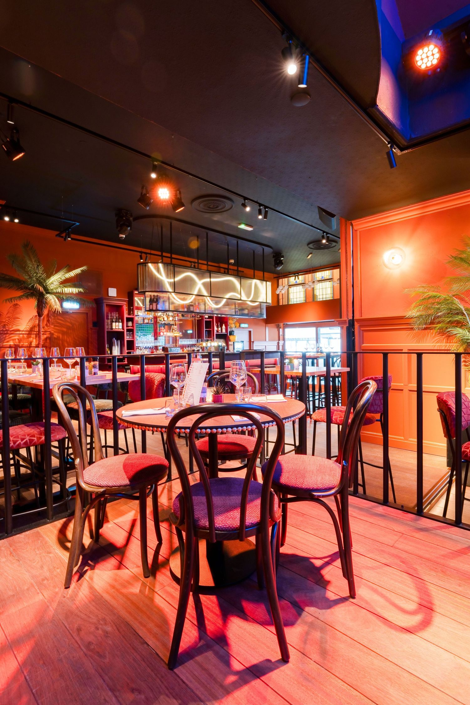 Cozy bar interior with round tables, wooden chairs, red lighting, neon signs, and a warm ambiance.