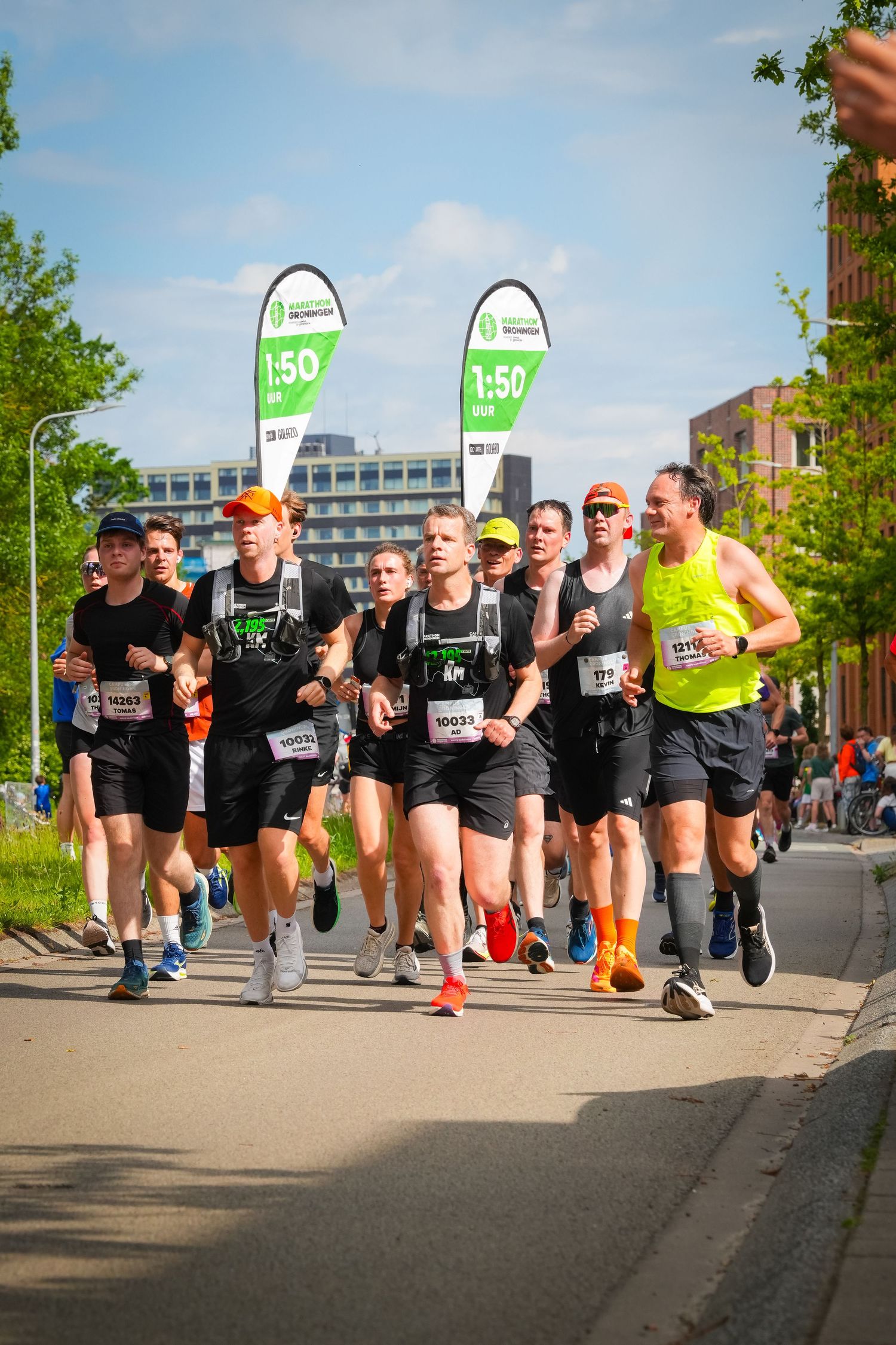 Group of runners competing in a road marathon race, wearing numbered bibs, with pace banners visible in the background.