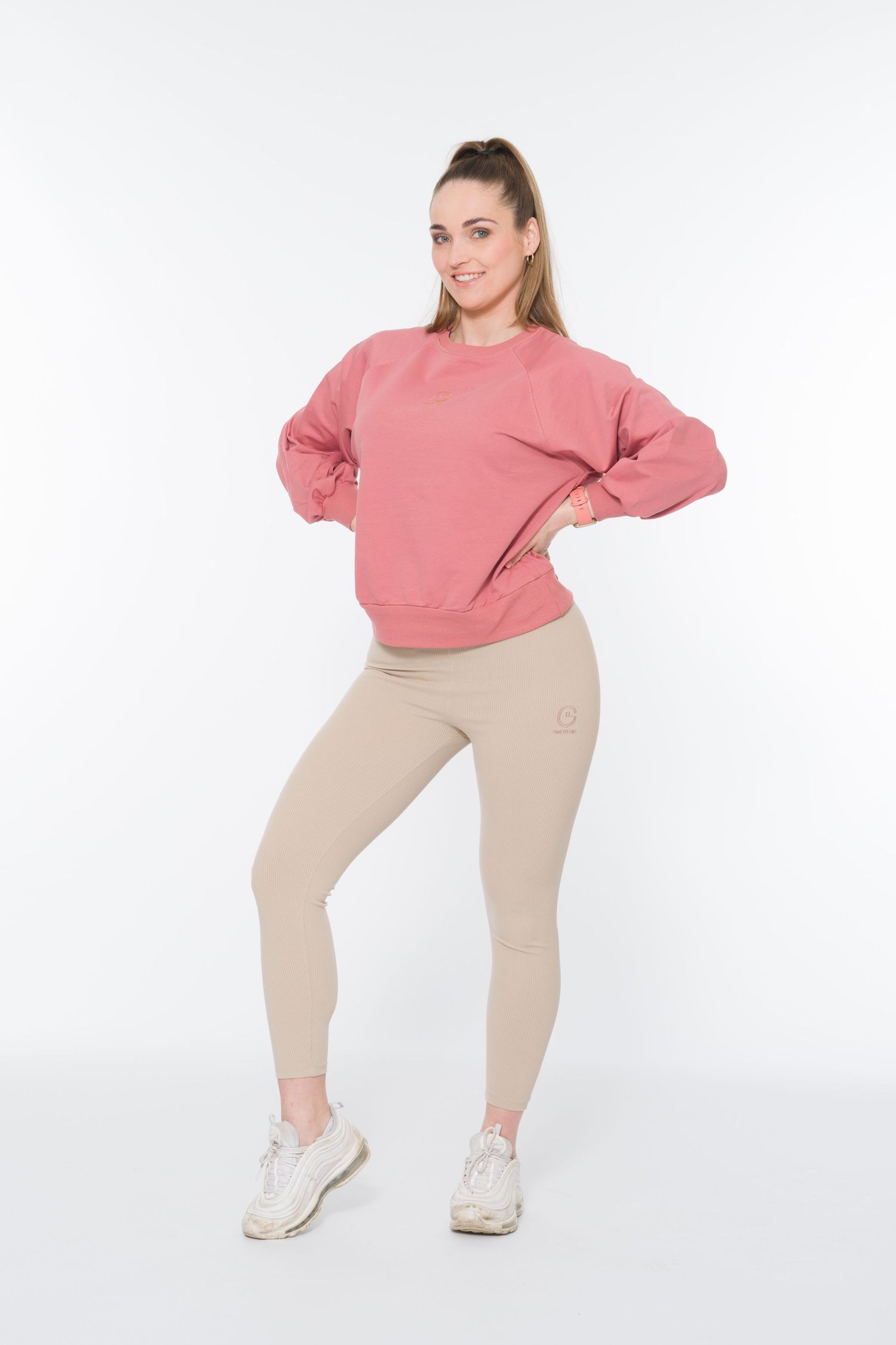 Woman in pink sweatshirt and beige leggings posing against white background.