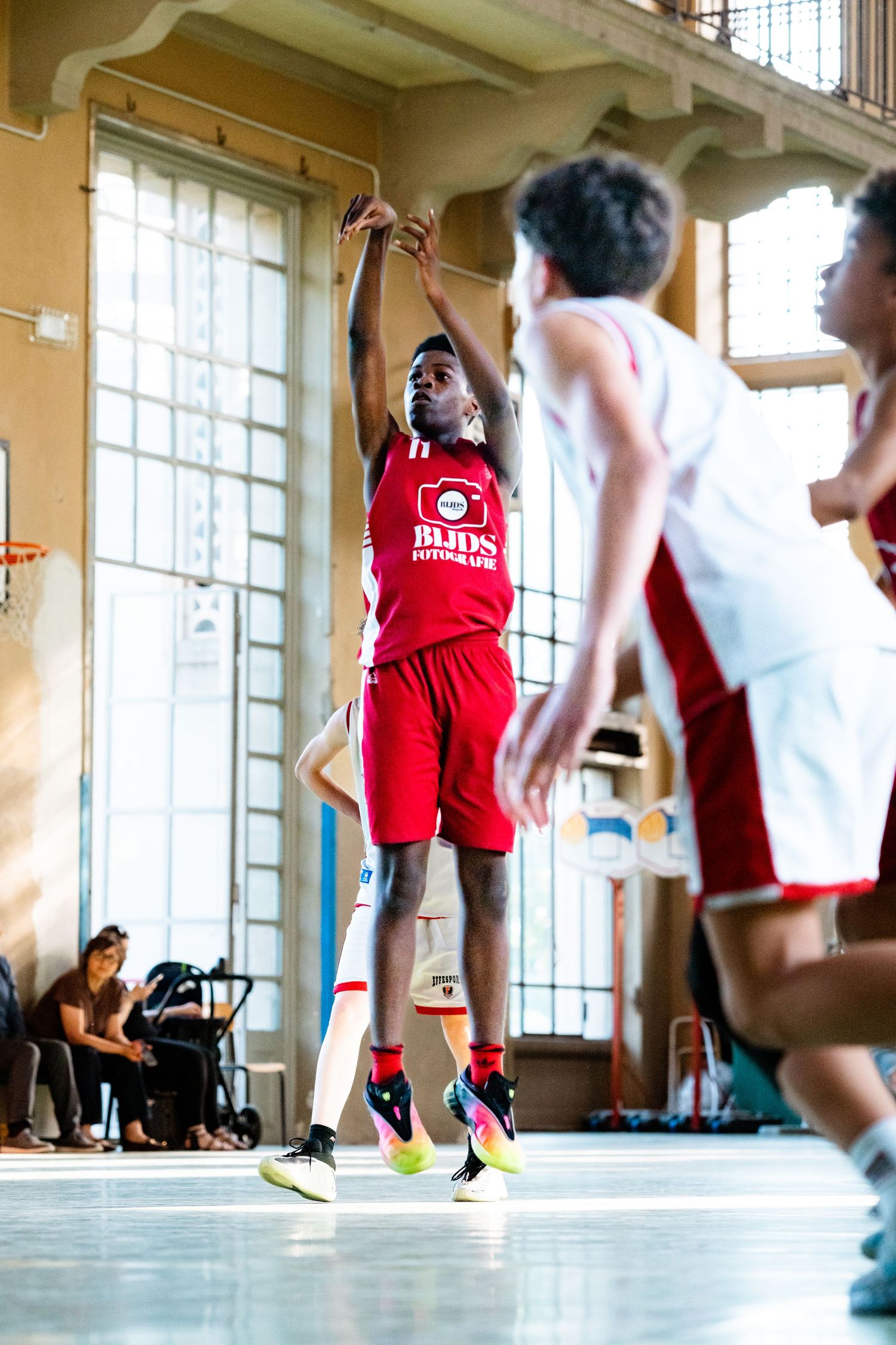 Young basketball player in red uniform jumps to shoot during an indoor game while defenders attempt to block.
