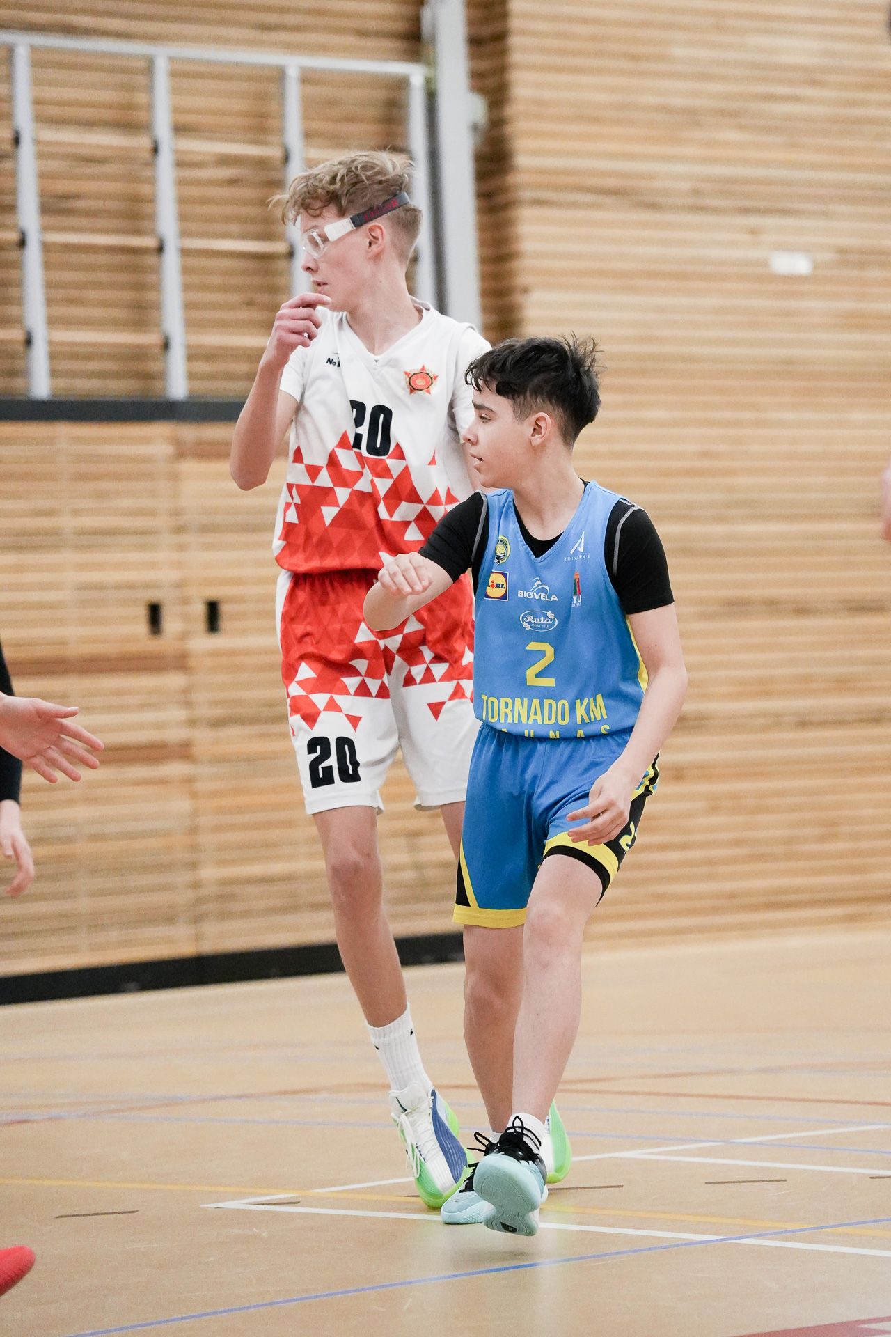 Two young basketball players competing on an indoor court, one wearing a blue Tornado kit number 2, the other in white number 20.