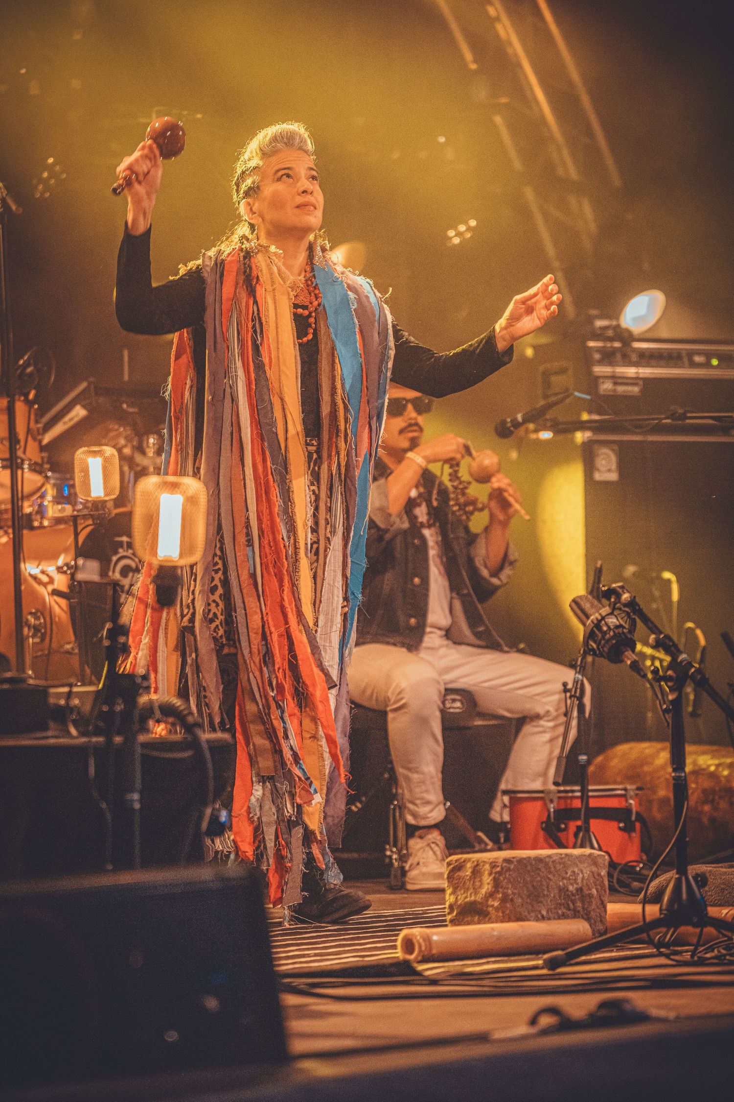 A performer in colorful tasseled costume dances energetically on stage during a live concert performance.