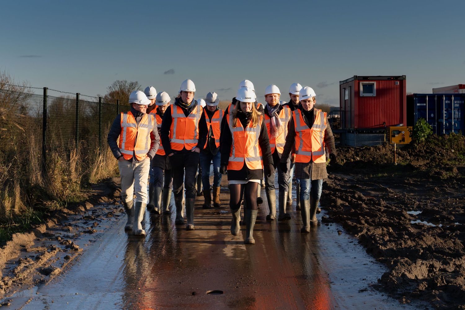 Group of construction professionals in orange vests and white hard hats walking on a muddy site.