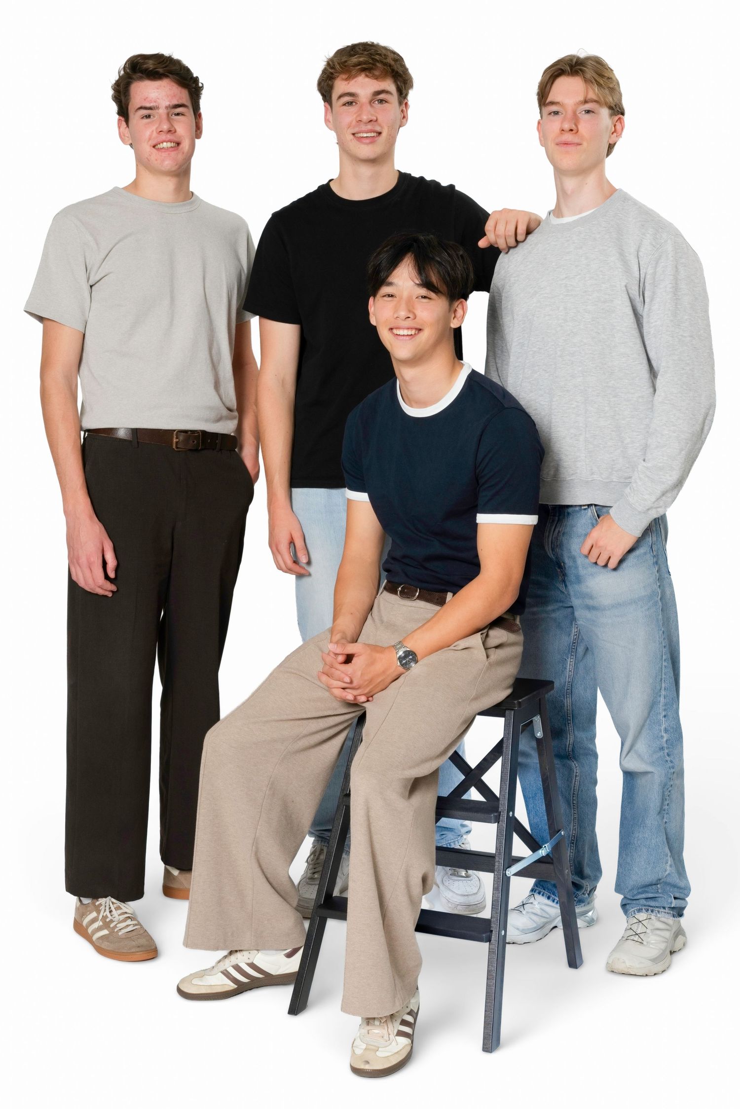 Four smiling young men in casual outfits pose together, three standing and one seated on a black wooden stool, against a white background.