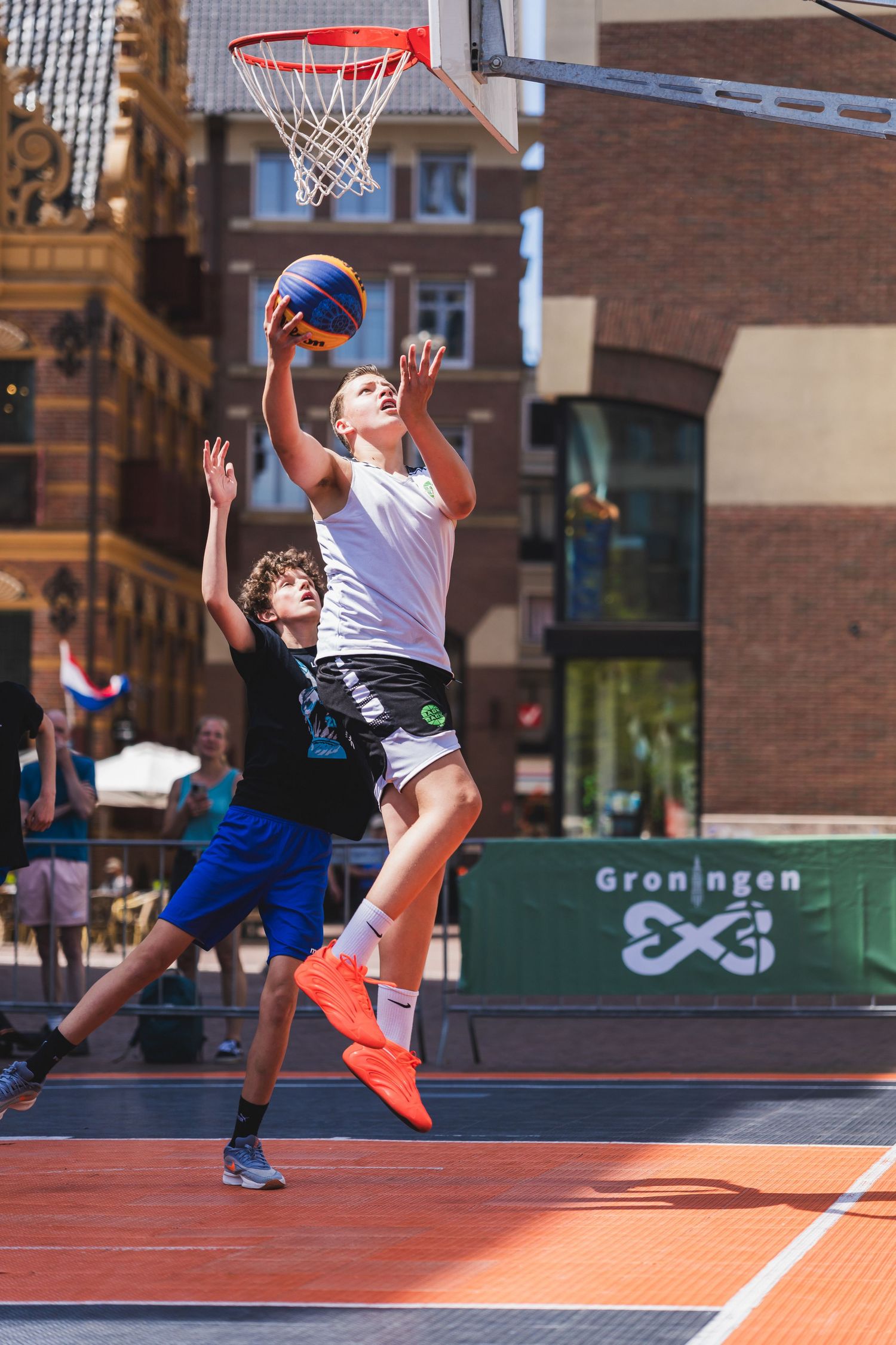 Two young players compete in a 3x3 street basketball game in Groningen, with one player leaping to score.