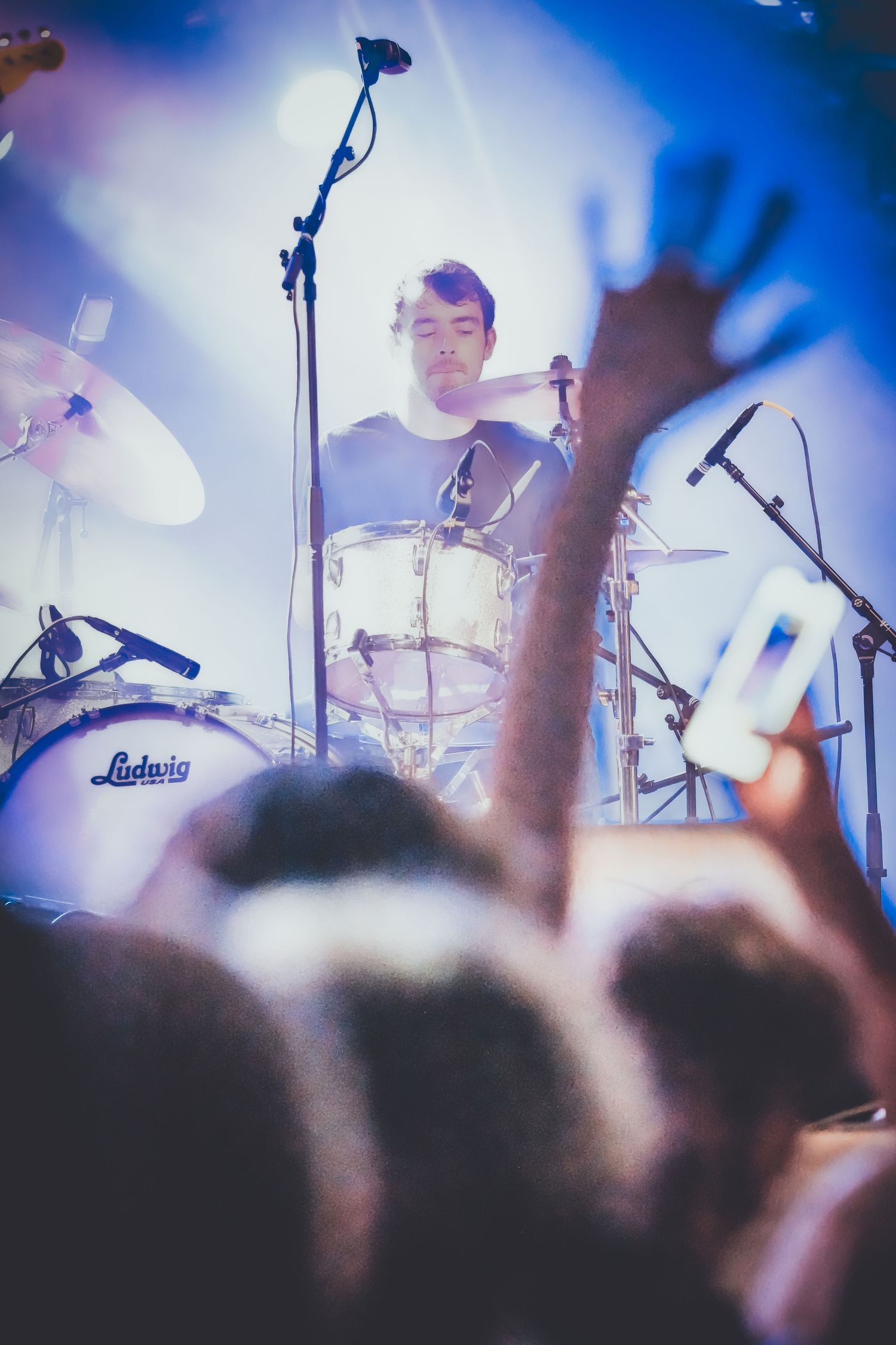 A drummer performs on stage with a Ludwig drum kit while silhouetted crowd members watch at a live concert.