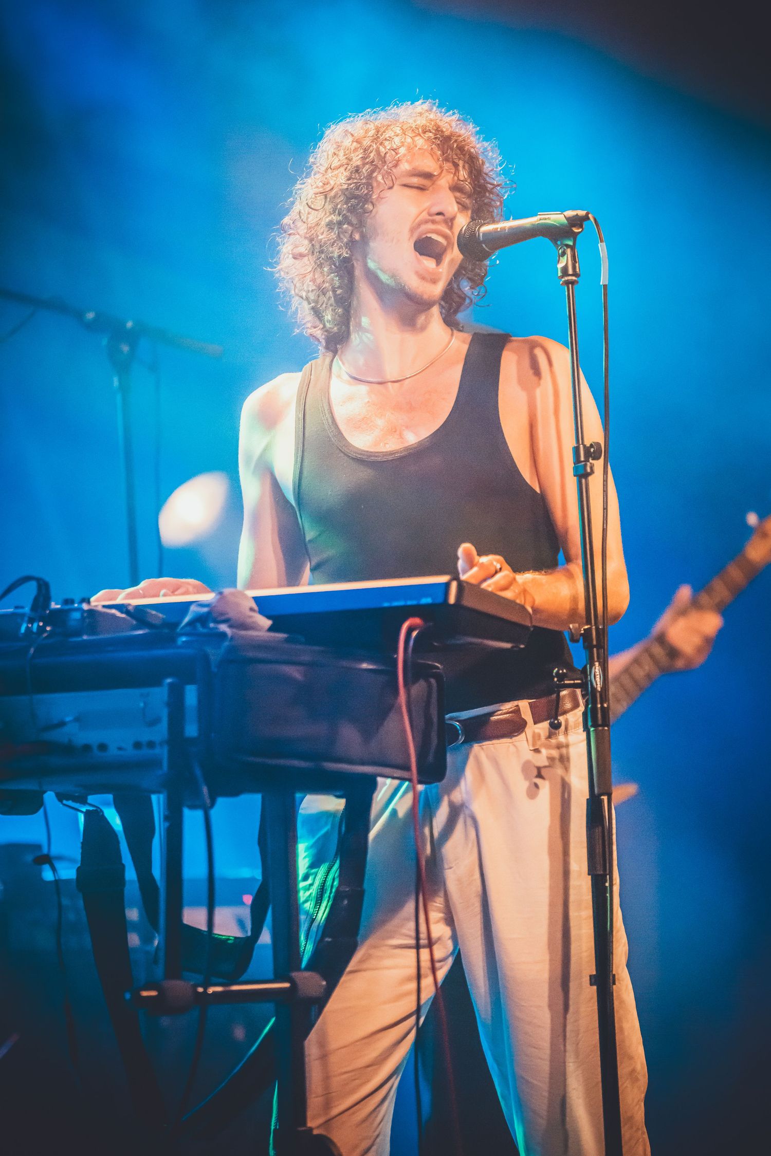Musician performing passionately at keyboard while singing into microphone on stage with dramatic blue lighting.