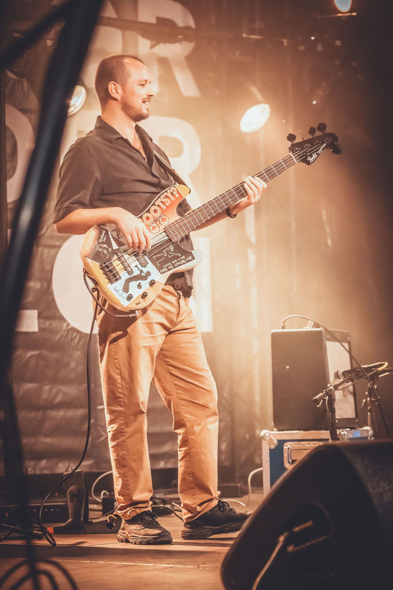 Bass guitarist performs on stage in khaki pants playing a dark-colored bass guitar under warm stage lighting.