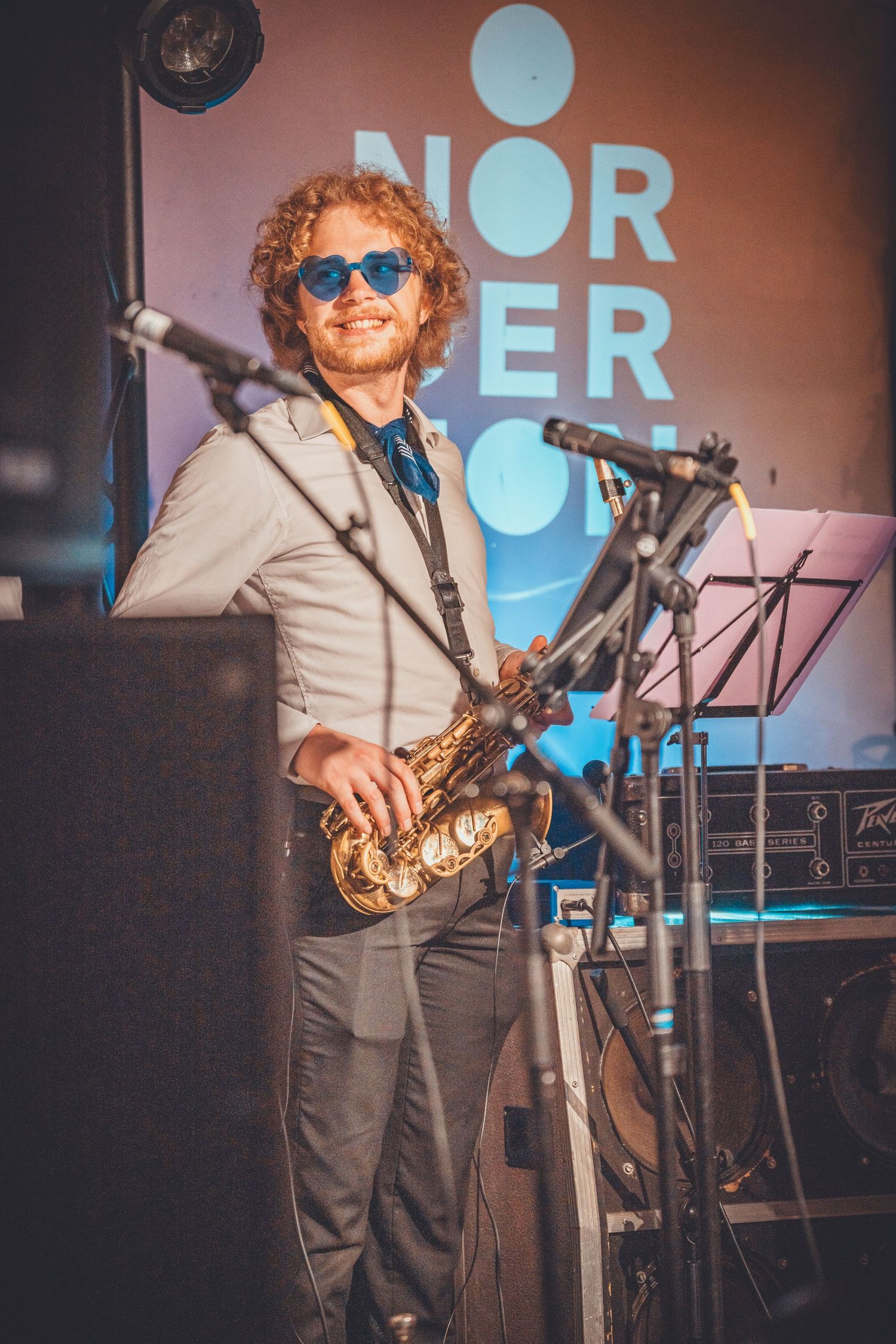 Musician plays saxophone on stage wearing formal attire with promotional backdrop.