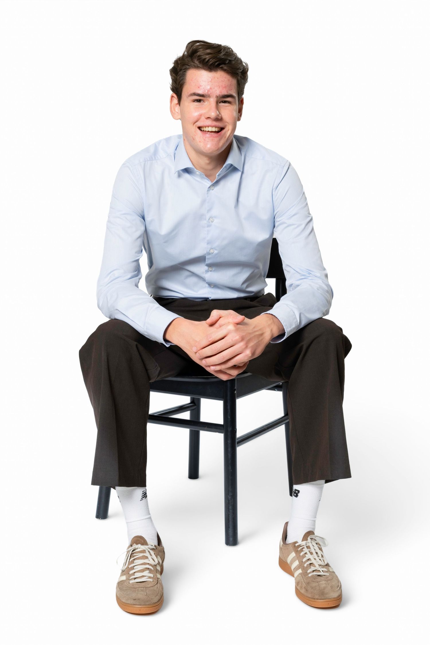 Smiling young man in a light blue shirt, dark trousers and tan sneakers sits on a black wooden chair with his hands clasped.