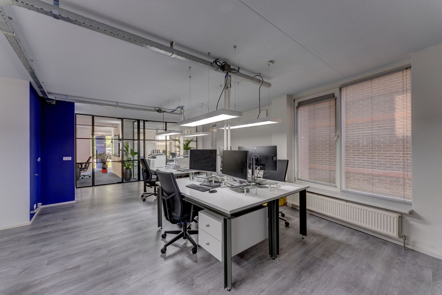 Modern open office workspace with gray floors, white desks, black chairs and blue accent wall in an industrial style setting.