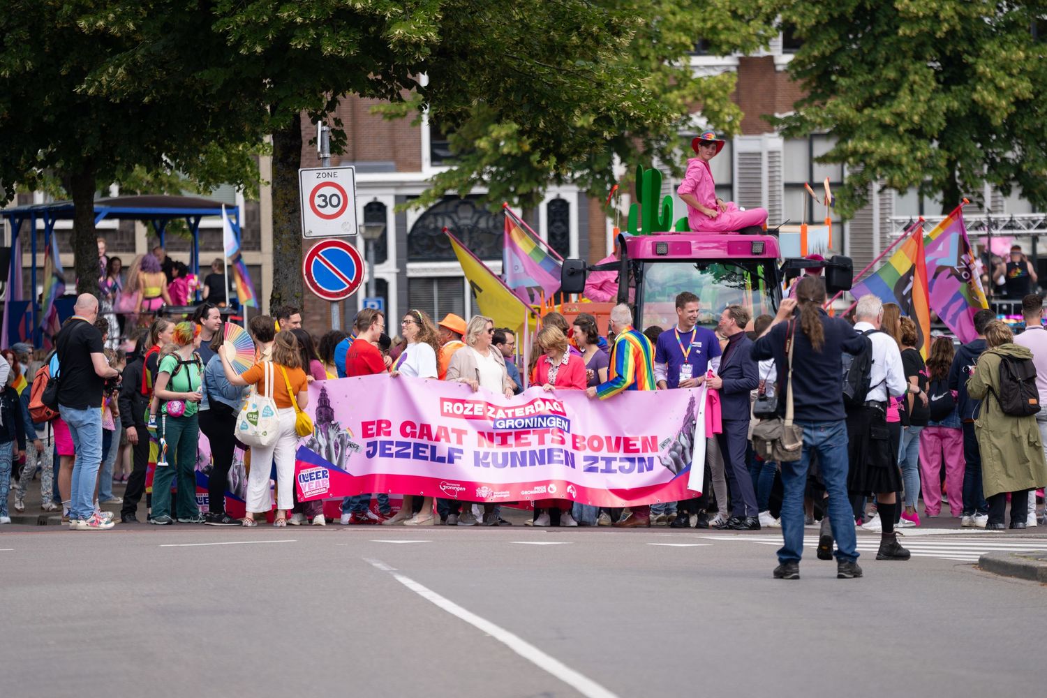 Pride parade participants march down a city street holding a bright pink banner while onlookers gather along the sidewalk.