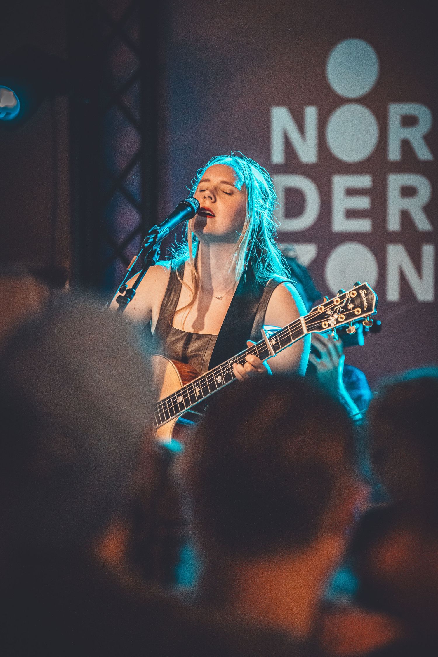 Performer with electric guitar singing into microphone on stage illuminated by blue lighting.