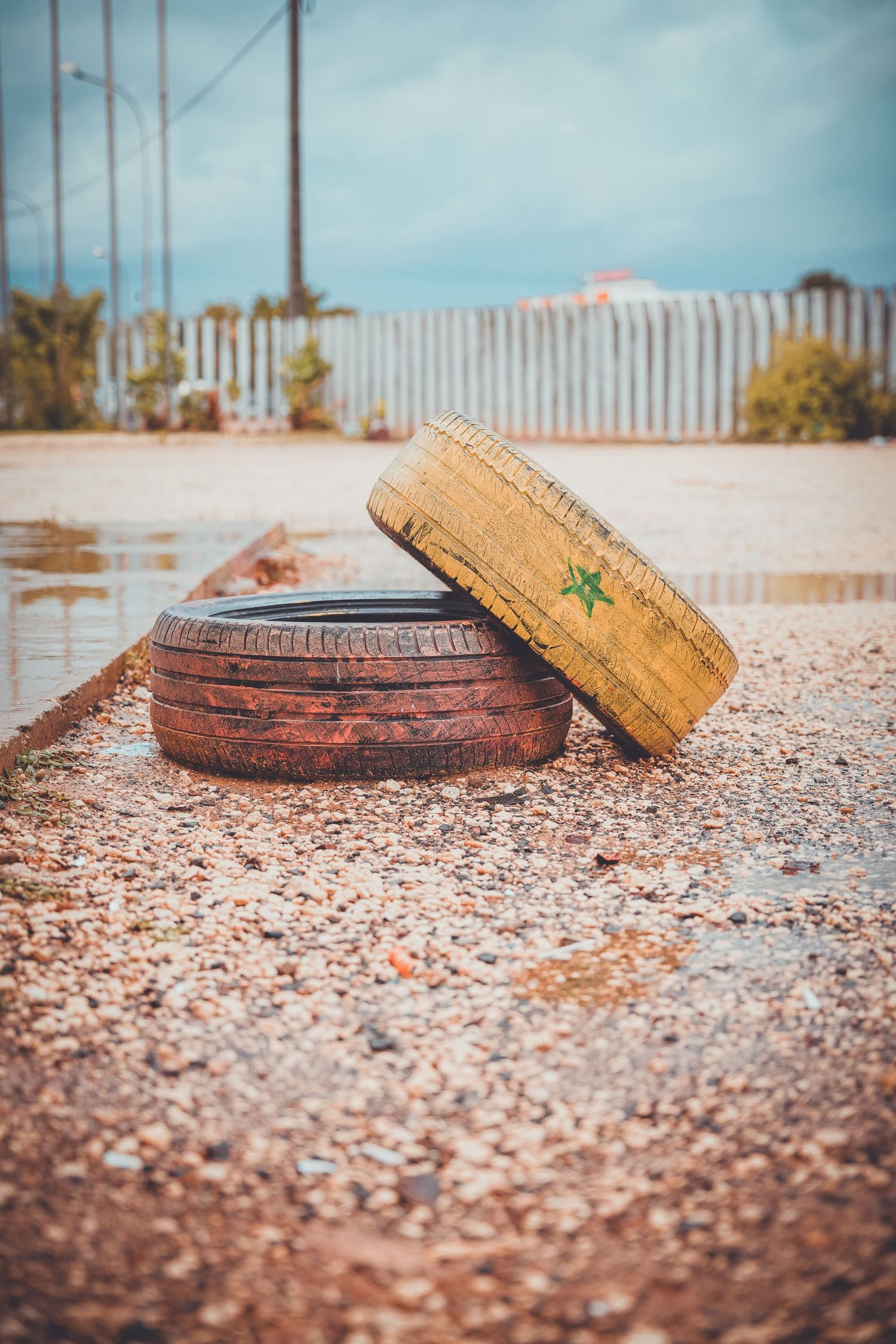 Two old worn tires stacked on gravel near a white fence under a cloudy sky.