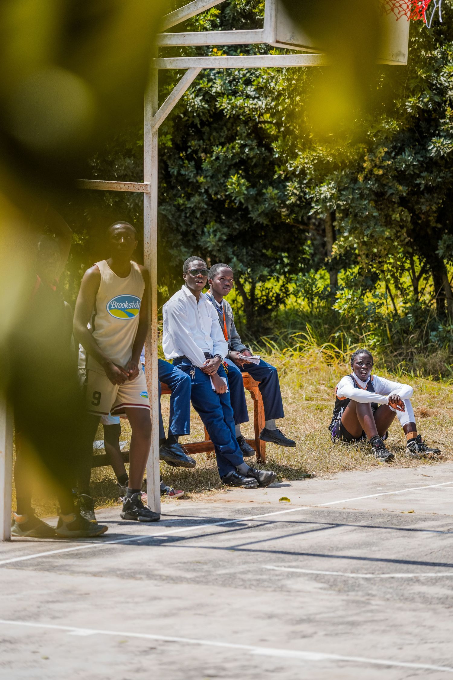 Men relaxing courtside at an outdoor basketball court surrounded by lush green trees in Africa.
