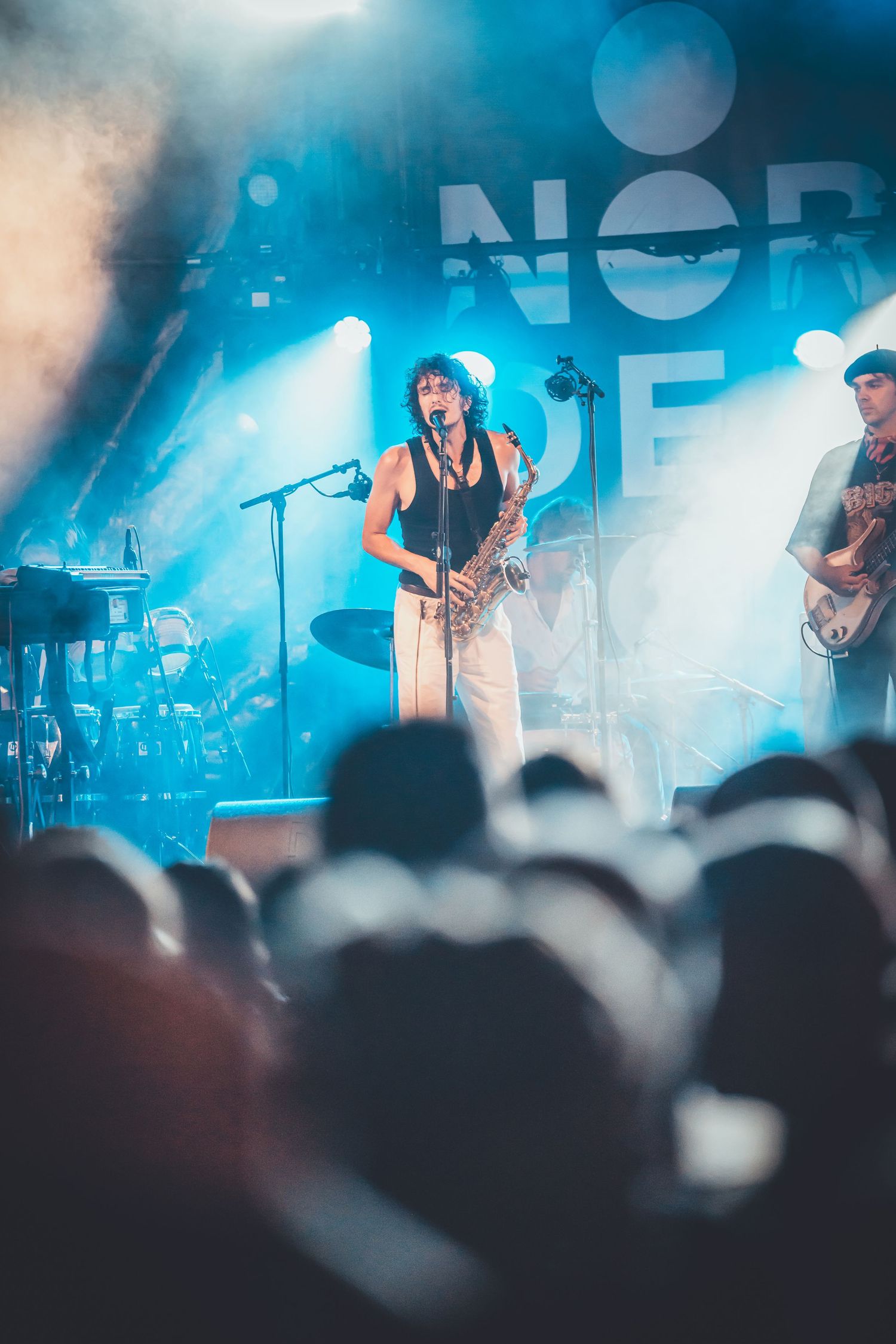Live concert performance with stage backlit in blue showing band members and silhouetted crowd in foreground.