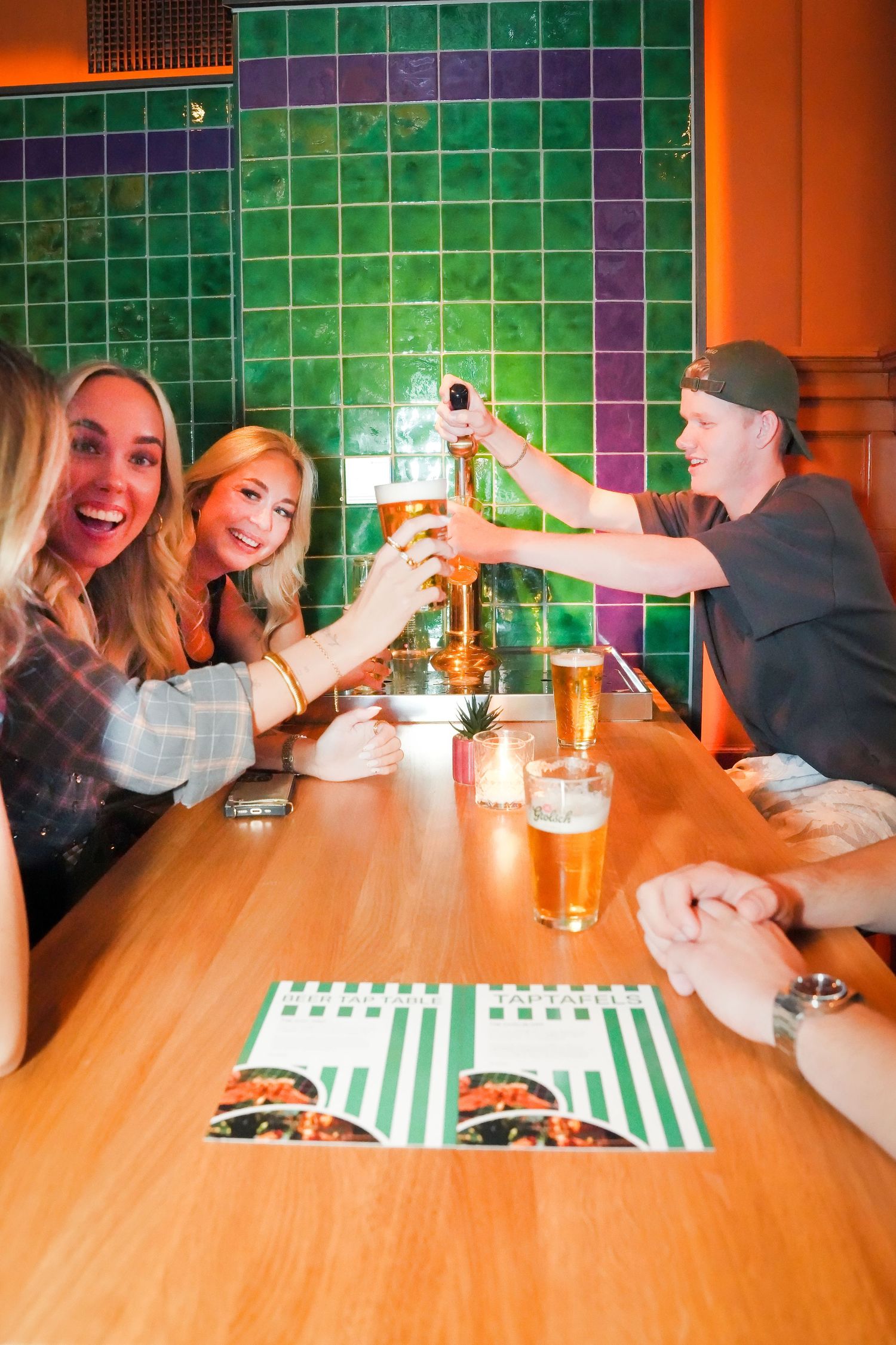 Friends toasting with beer glasses at a restaurant table with green tiles and menus.