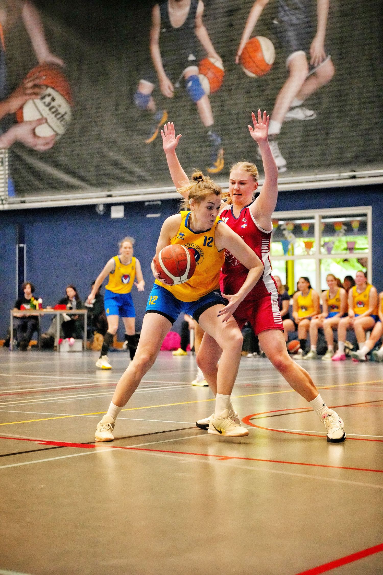 Female basketball player in yellow jersey dribbling against red-uniformed defender in indoor court game.