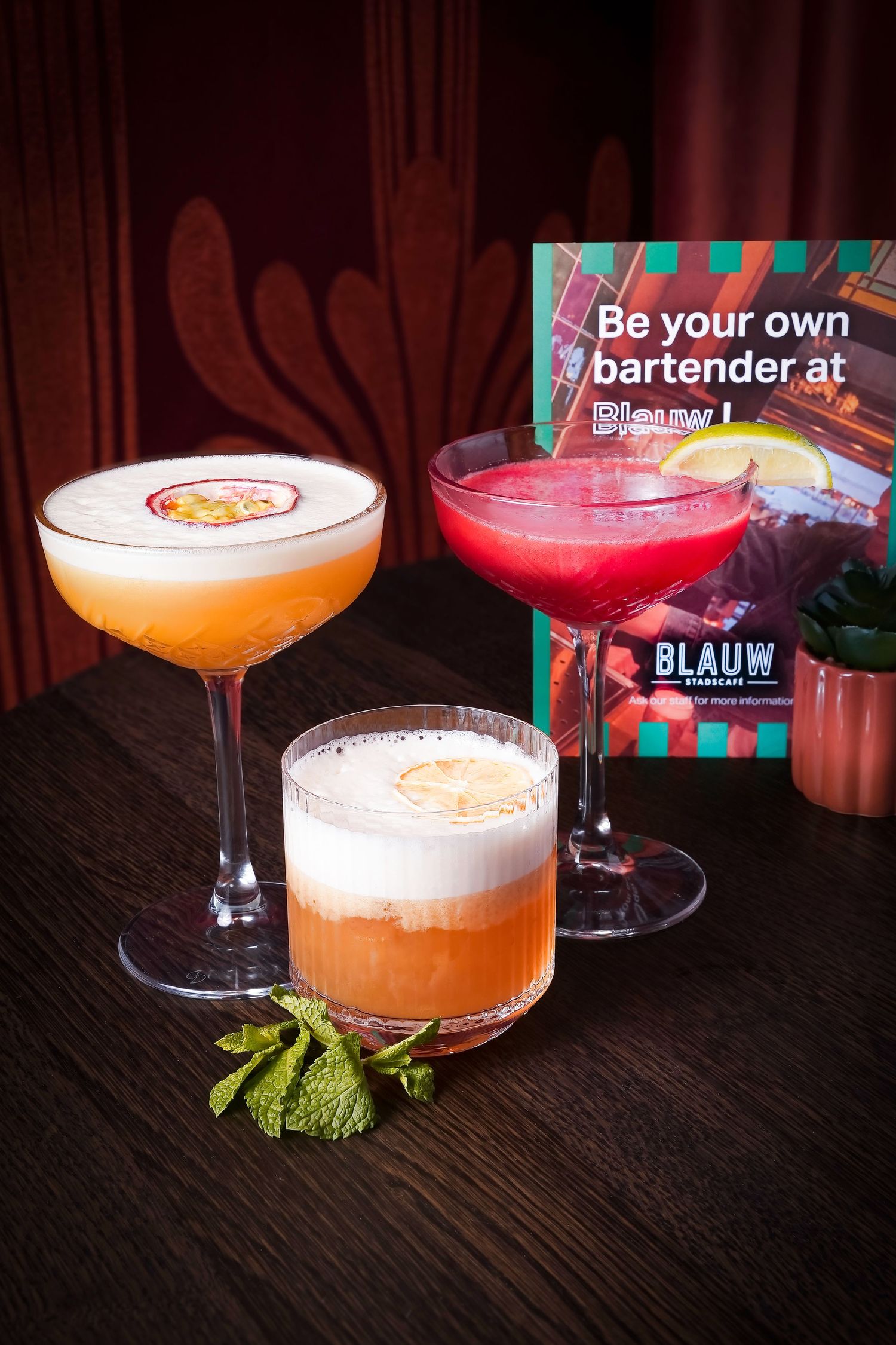 Three colorful cocktails displayed on a dark wooden bar next to a Blauw bartender experience promotional sign.