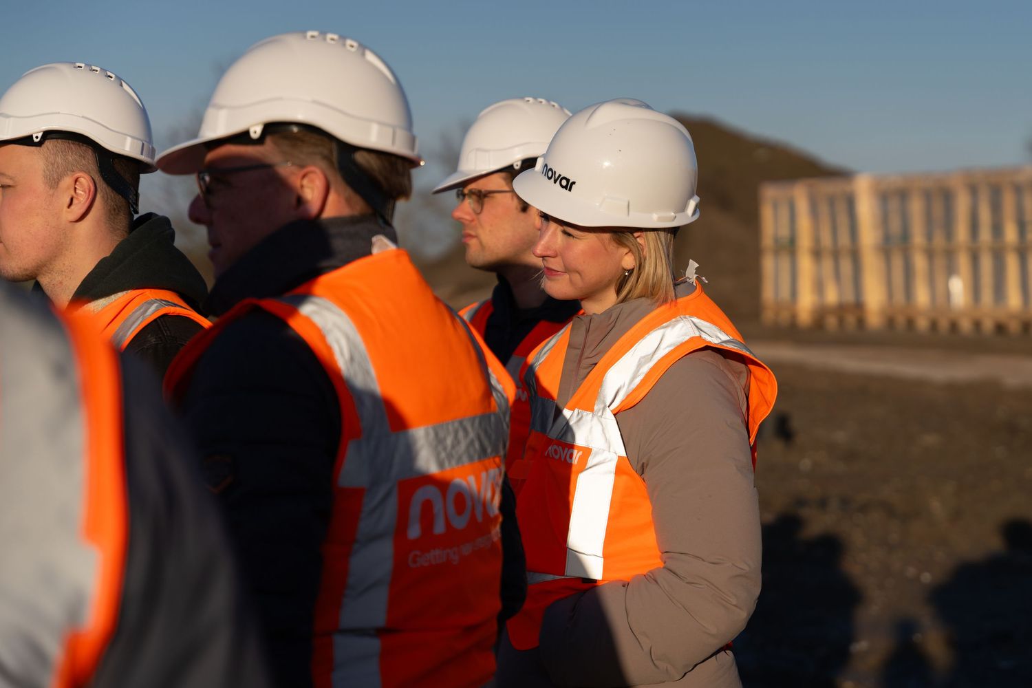 Construction workers wearing orange safety vests and white hard hats stand together at a worksite during sunset.