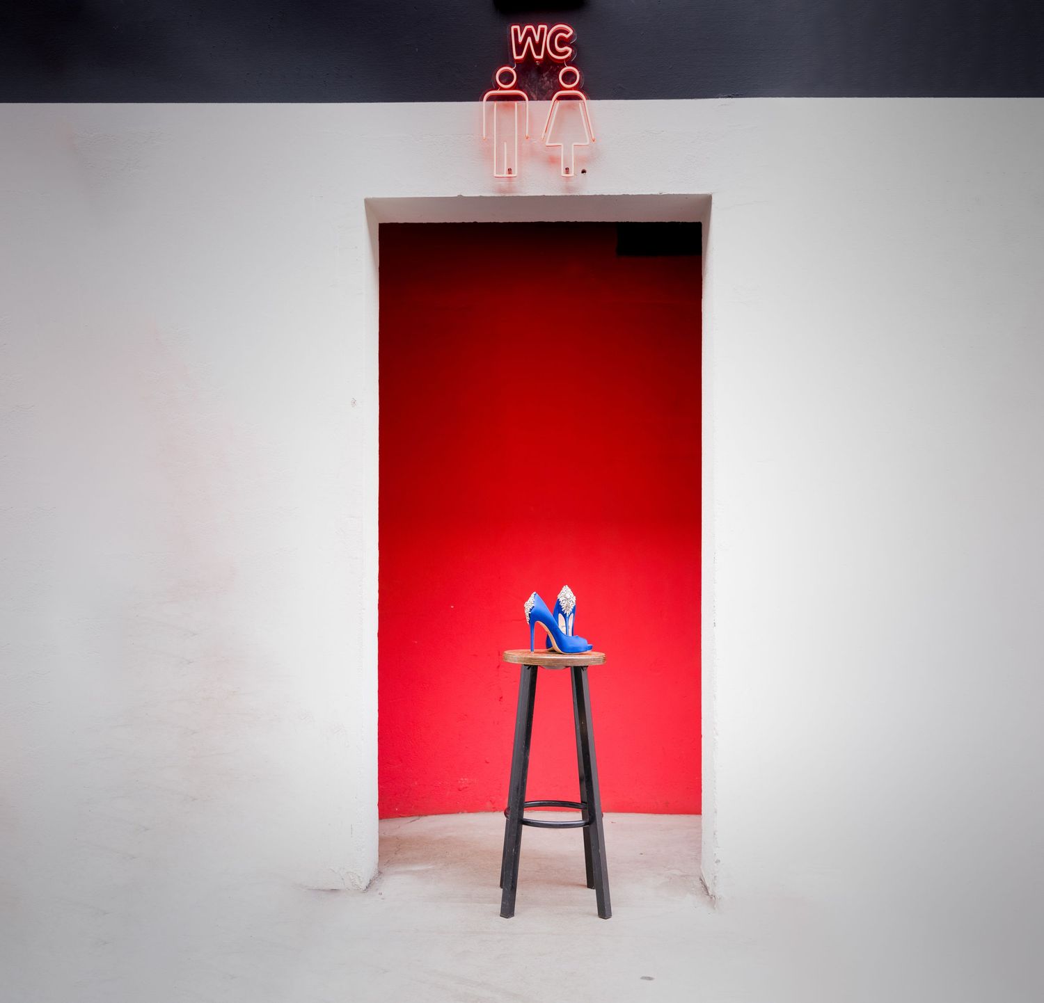 Minimalist scene of a blue item on tall stool against red wall with neon sign above.