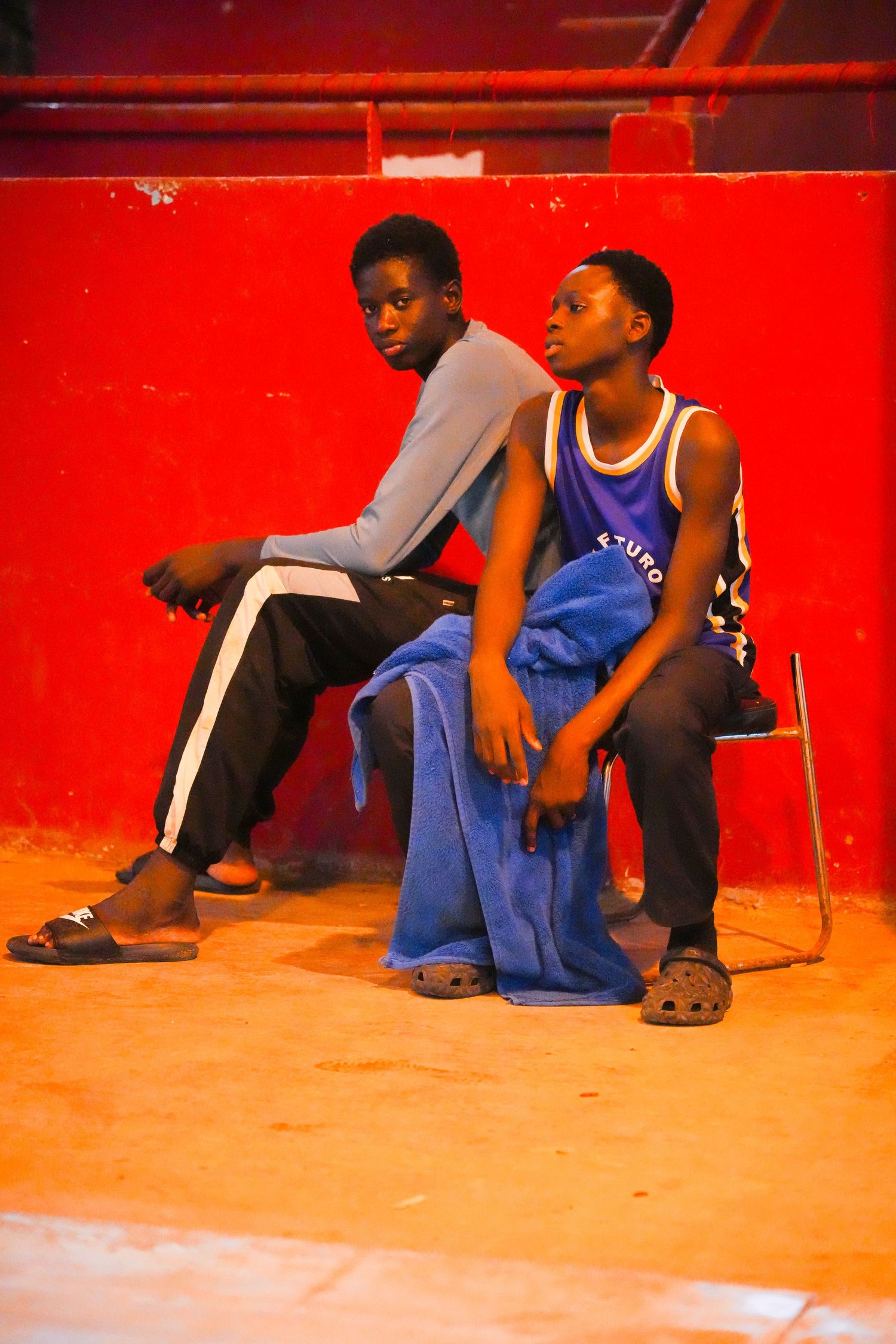 Two young African boys sitting against a vibrant red wall, one in a gray hoodie and the other in a purple jersey.