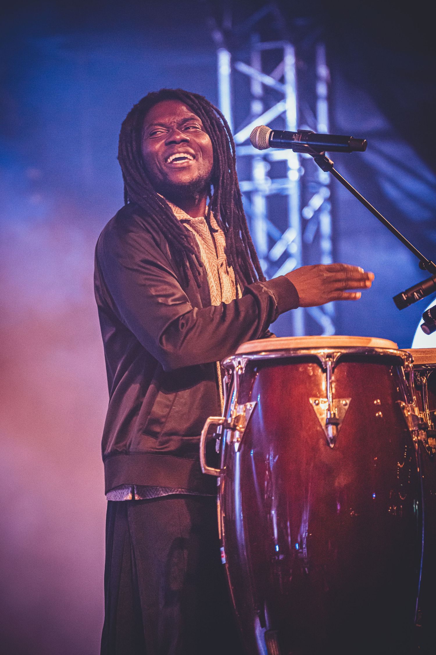 Energetic musician with dreadlocks performs on congas at a concert with stage lighting and microphone setup.