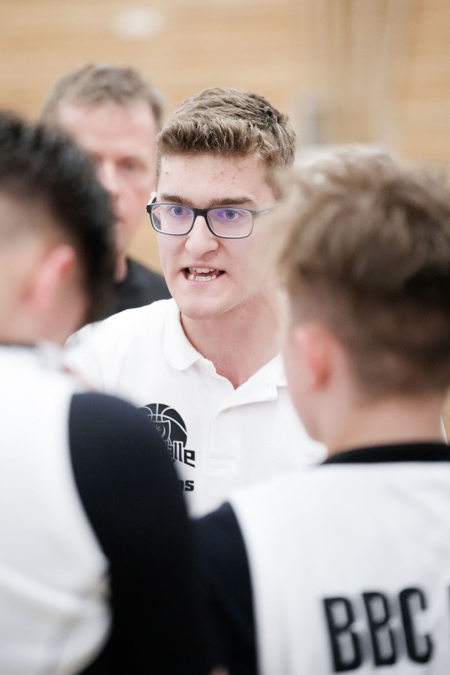 Young man in glasses and white BBC polo shirt speaking to teammates in a basketball team huddle.