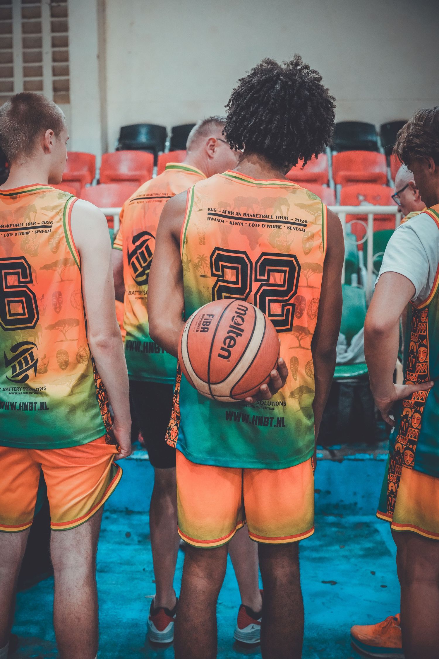 Basketball players in orange and green jerseys huddle together, with player 22 holding a Molten basketball during a pre-game moment.