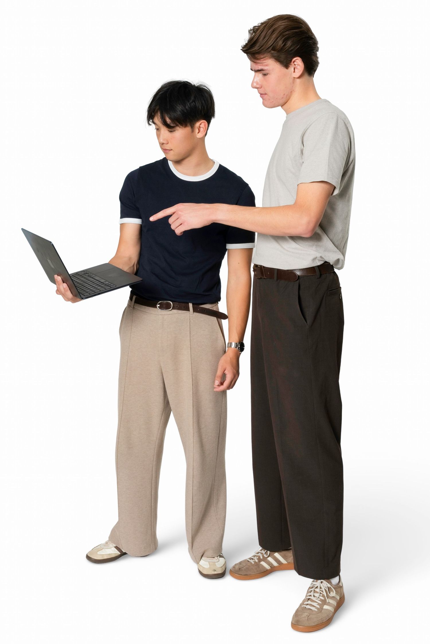 Two young men in casual outfits stand together while one holds a laptop and the other points at its screen.