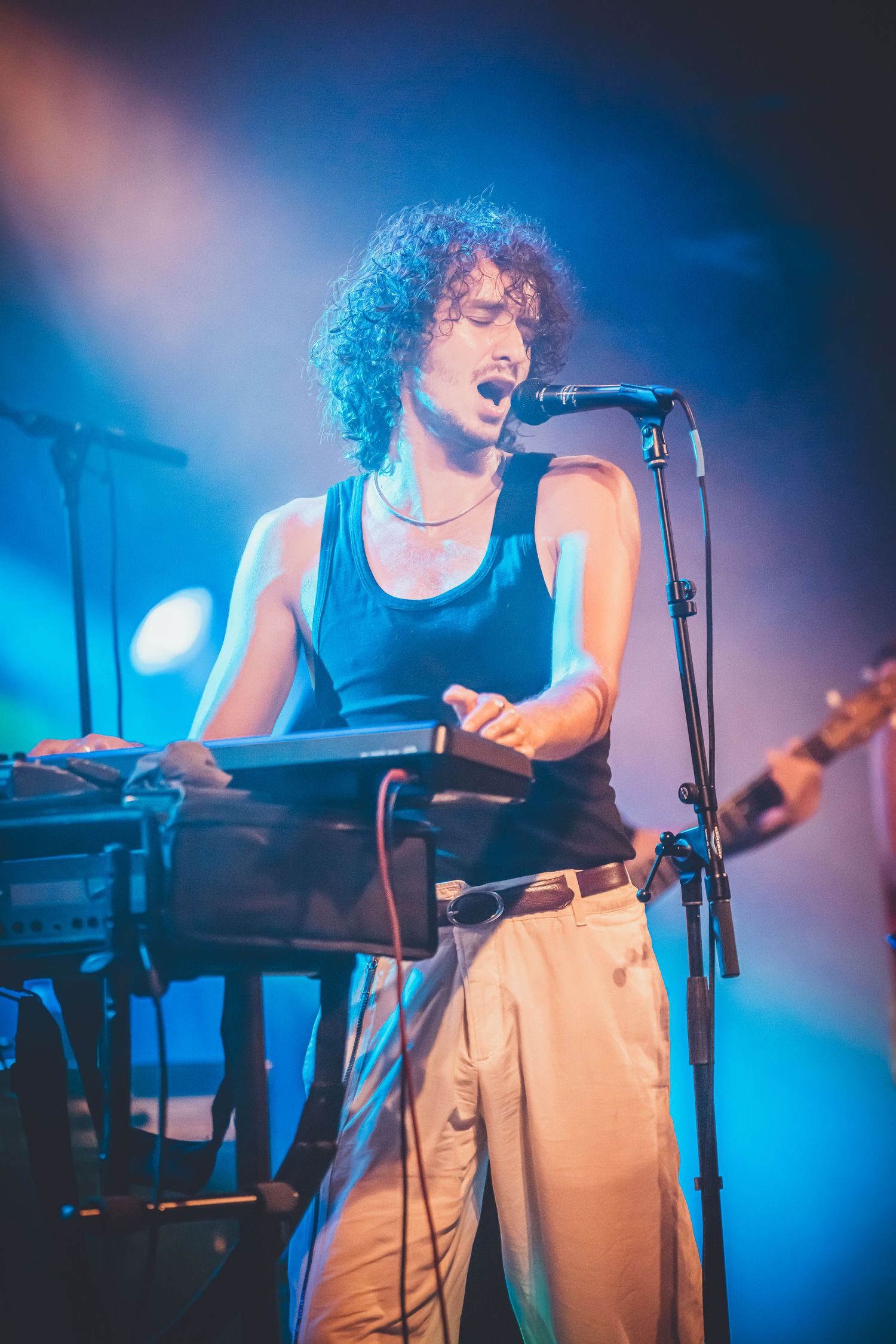 Performer in blue tank top and light pants singing passionately on stage with keyboard and microphone under blue lighting.