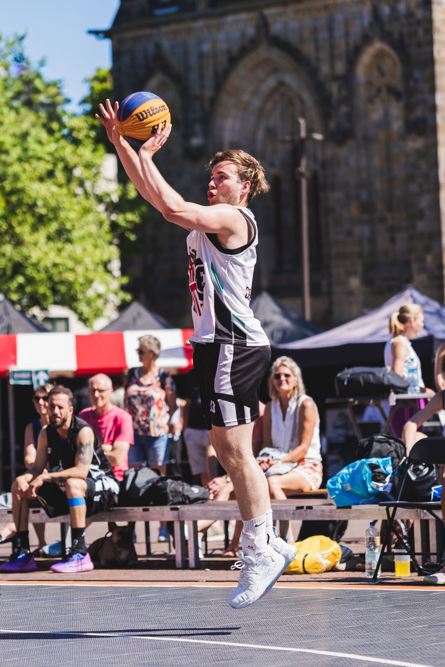Young man jumping to shoot a Wilson basketball during an outdoor 3x3 street basketball game in a sunny urban plaza.